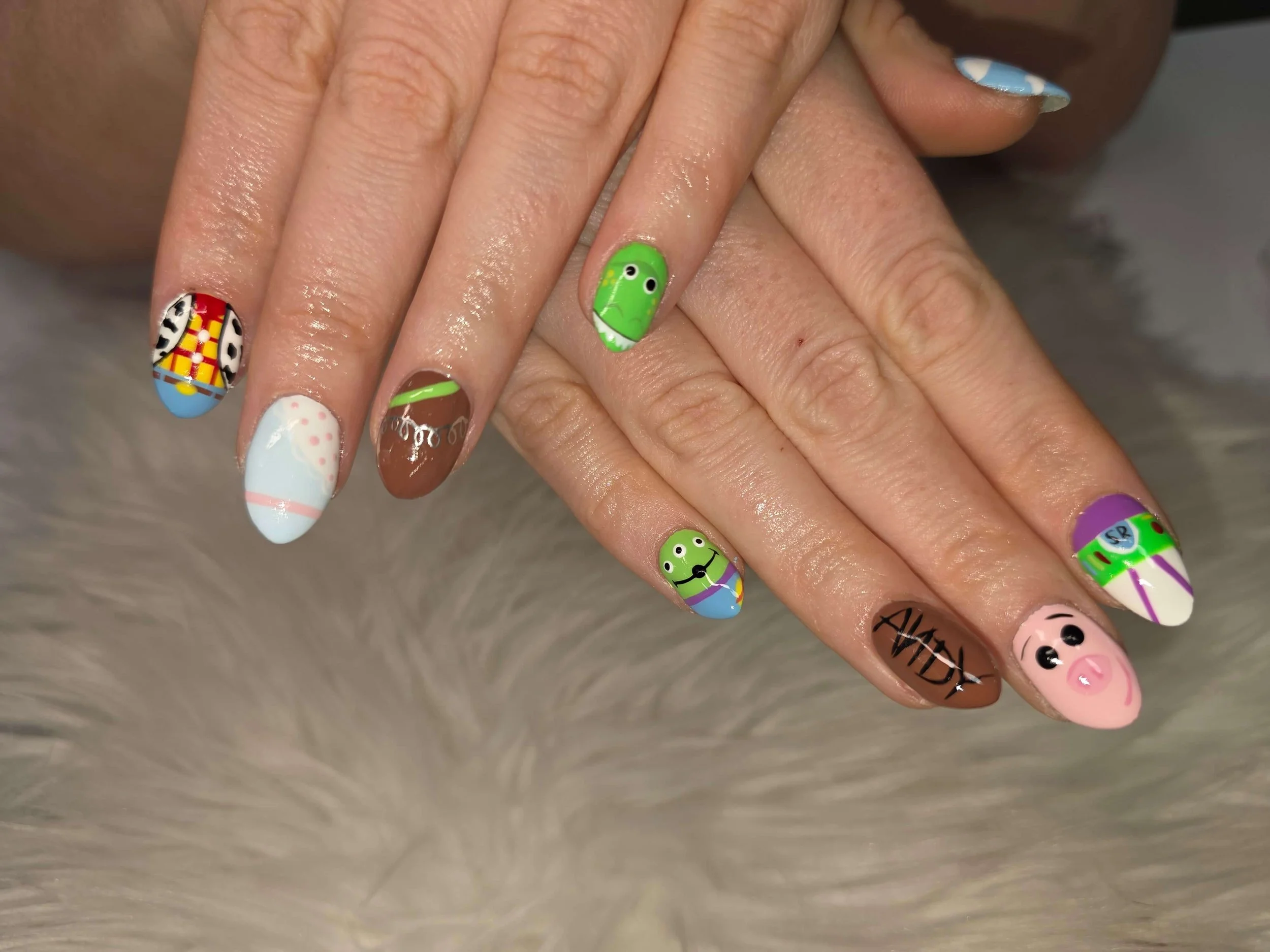 Close-up of a person's hand with colorful, animal-themed painted nails, including designs of a cow, pig, frog, cowgirl pattern, dinosaur, sheep, and a pastel pink and white pattern.