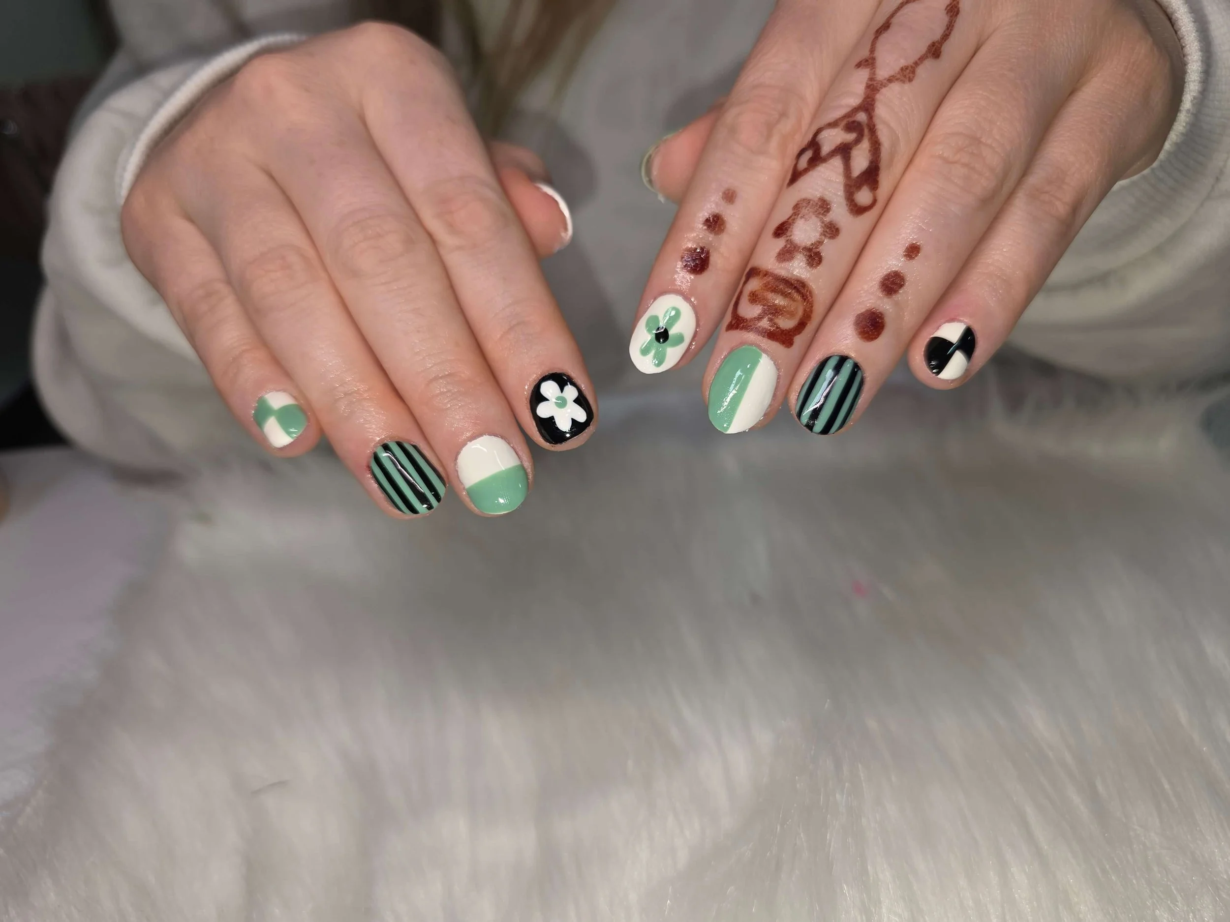 Close-up of a person's hand with decorated nails featuring black and white stripes, green and white designs, and flower patterns, along with henna tattoos on fingers.