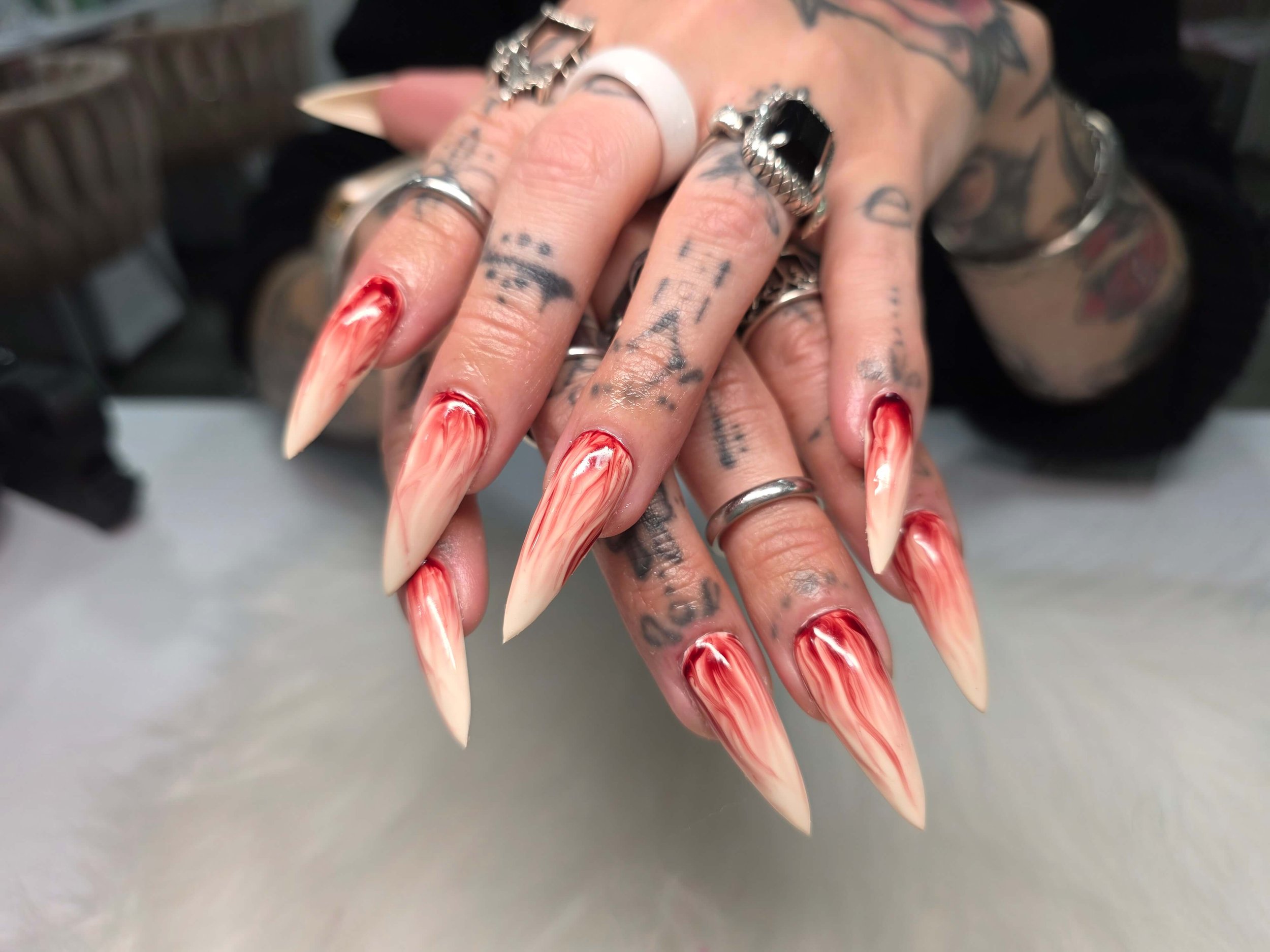 Close-up of multiple tattooed fingers with long, pointed, red and white marbled nails and various metallic rings.