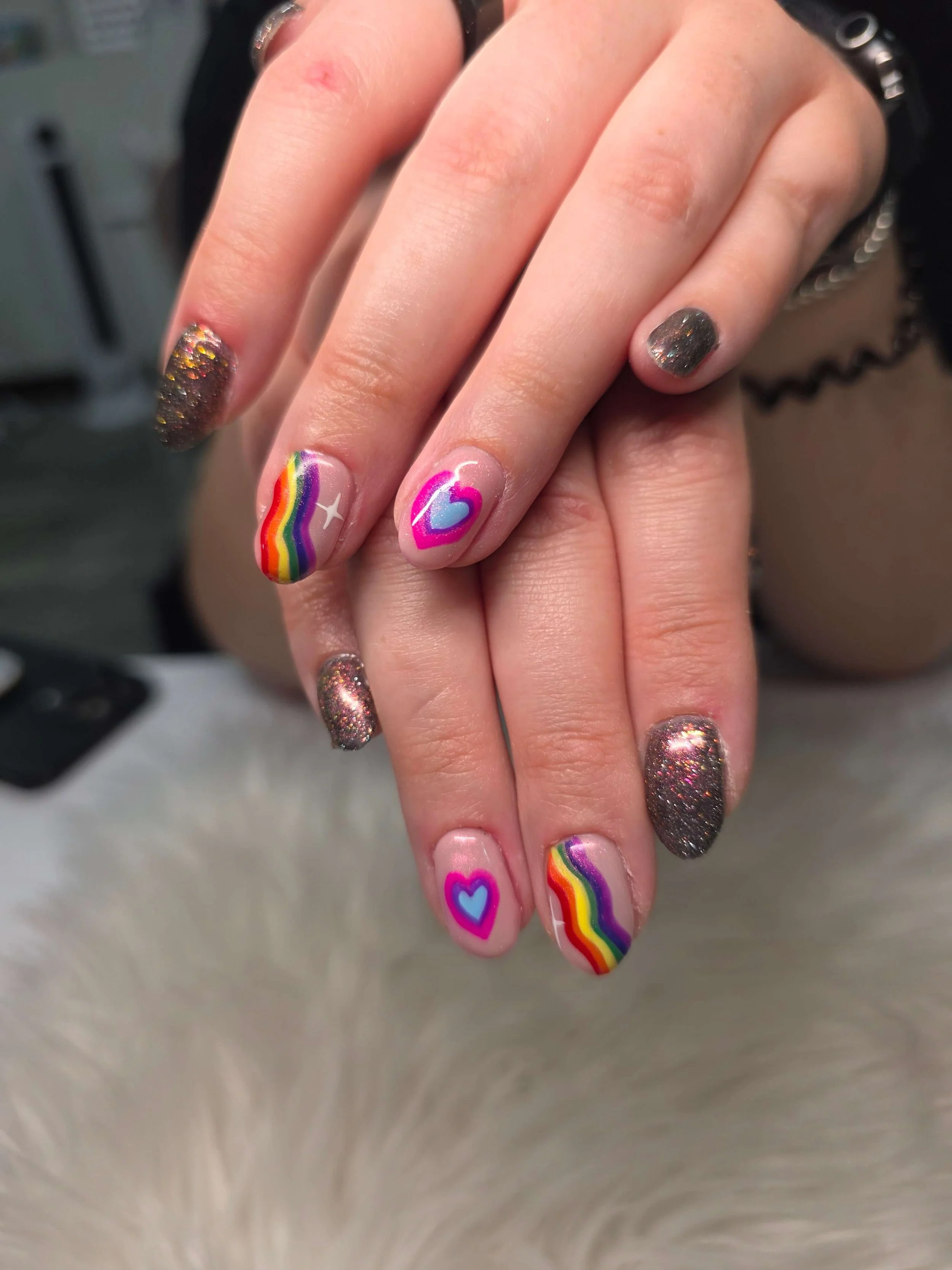 Close-up of a person's hands with rainbow and rainbow-themed nails, some with glitter and some with rainbow and heart designs.