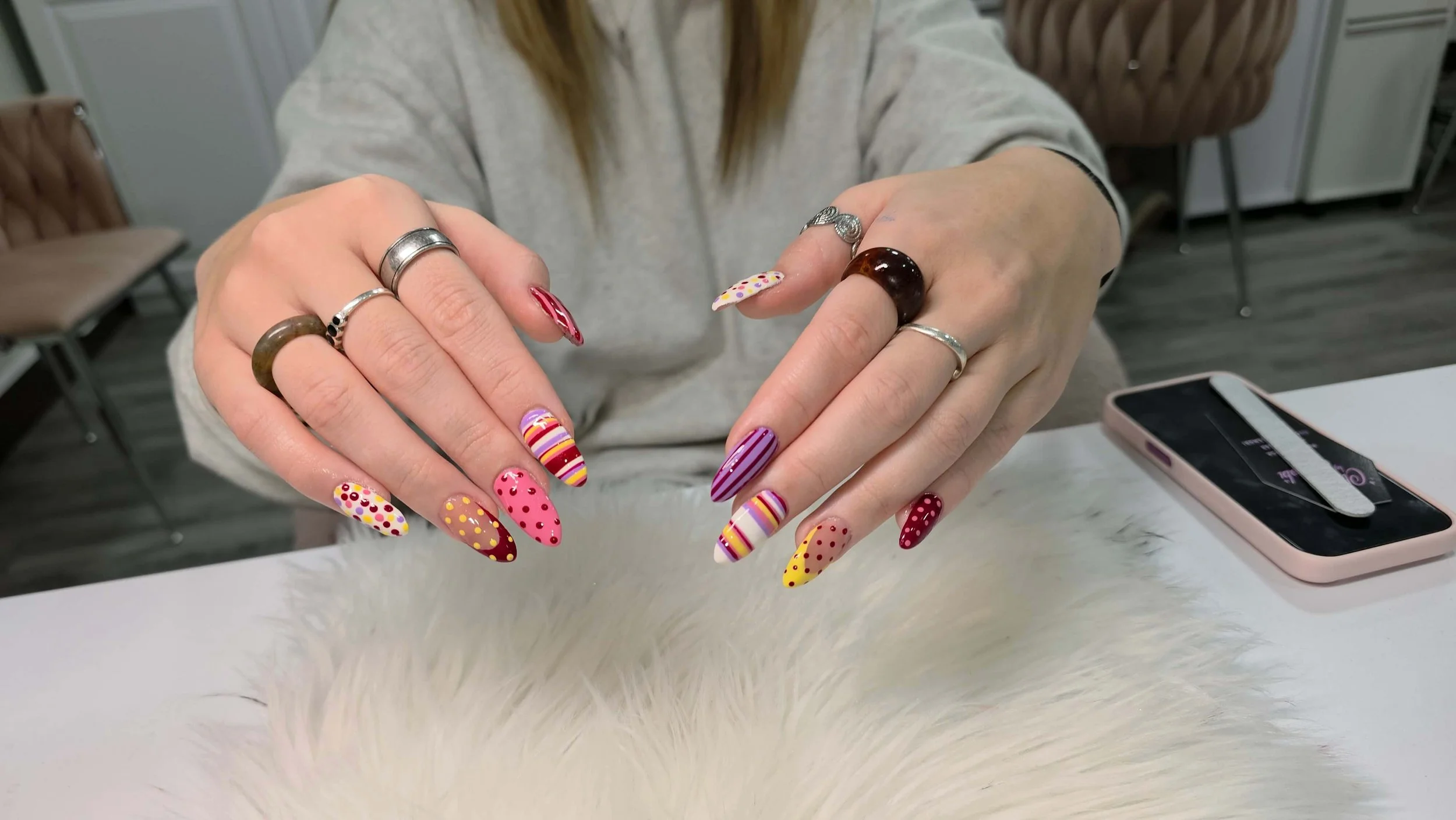 Person showing off colorful, patterned nail art with rings on fingers, sitting at a table with a smartphone nearby.