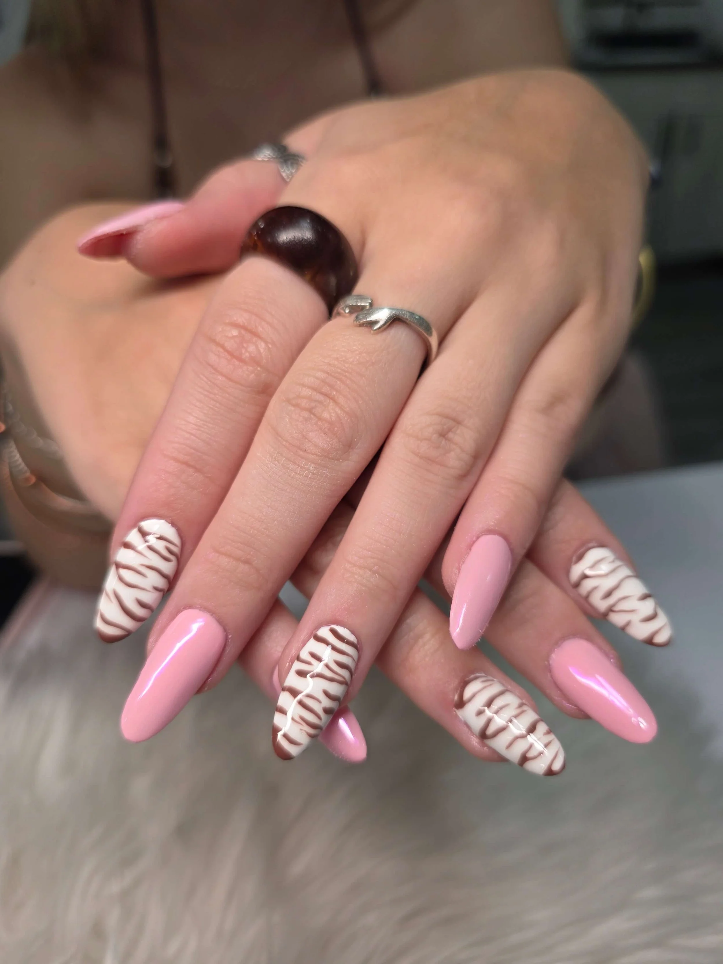 Close-up of hands with pink and white zigzag patterned manicured nails, wearing rings, with background of blurred hair and gray surface.
