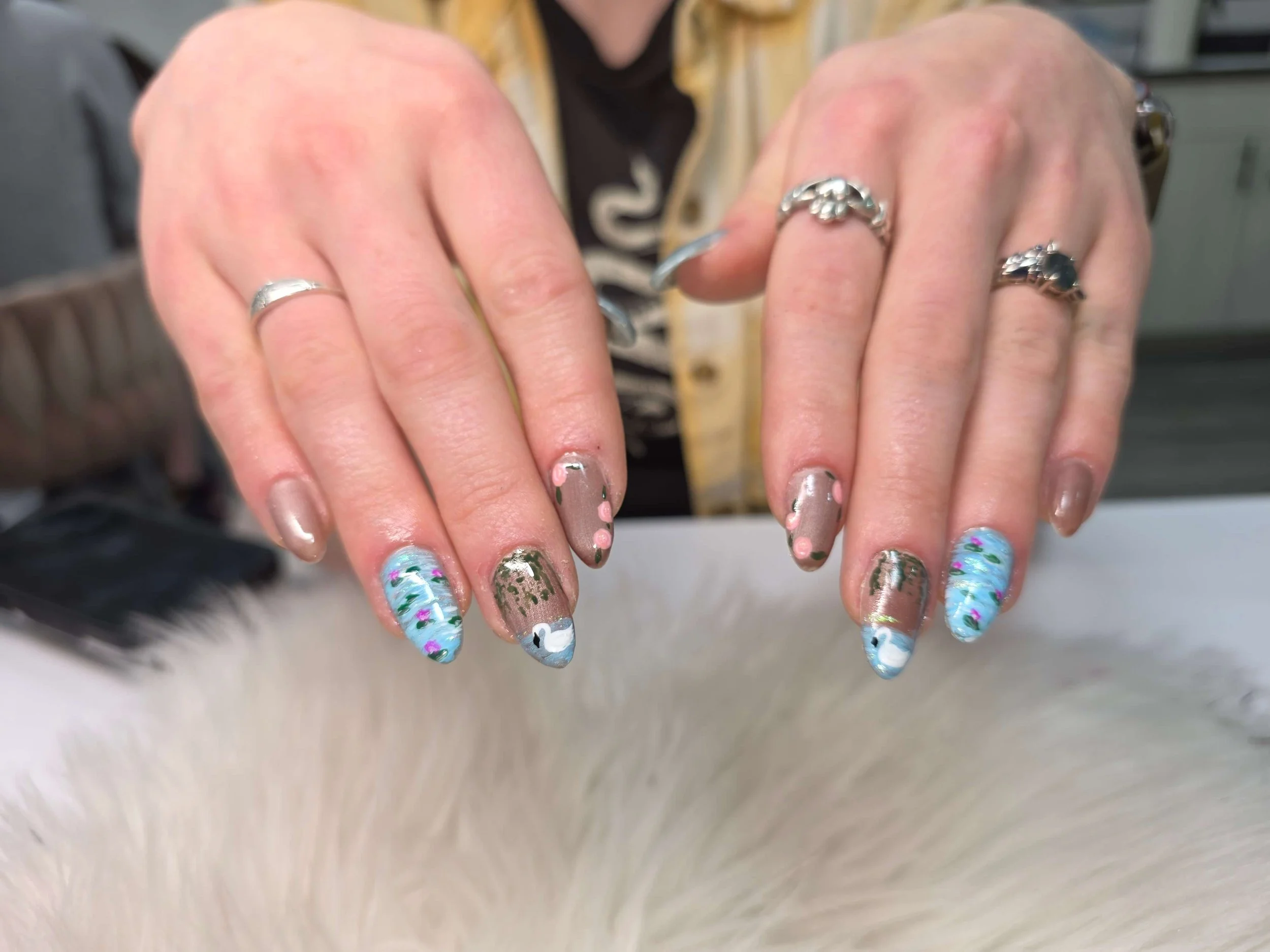 Close-up of hands with painted nails and silver rings, showing nail art with floral patterns, glitter, and abstract designs.