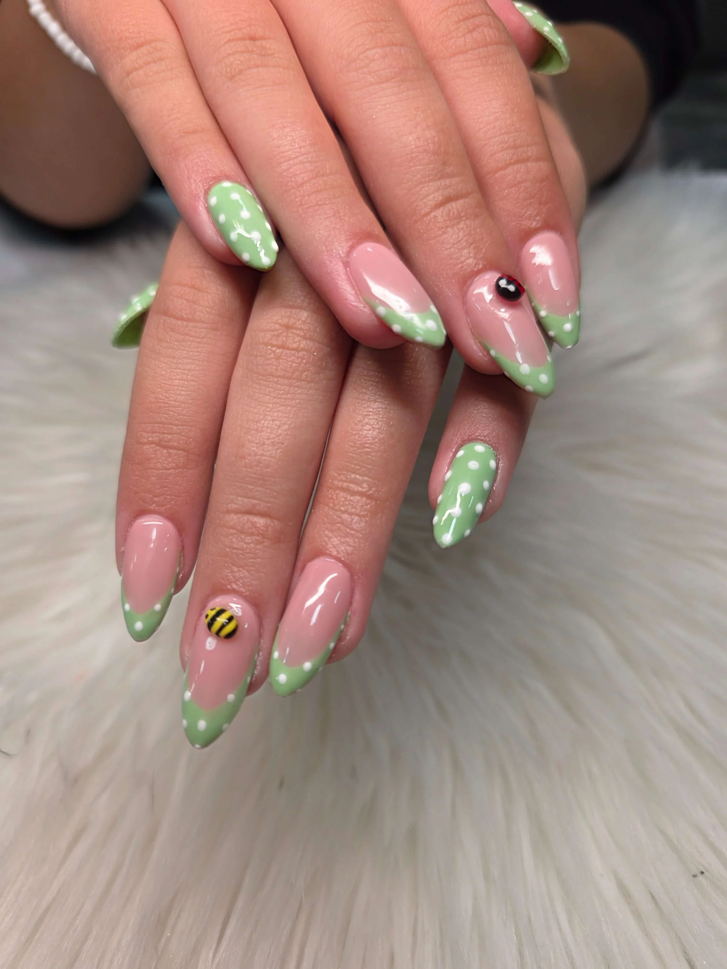 Close-up of hands with manicured nails painted in green with white polka dots, featuring a 3D bee design on one nail.