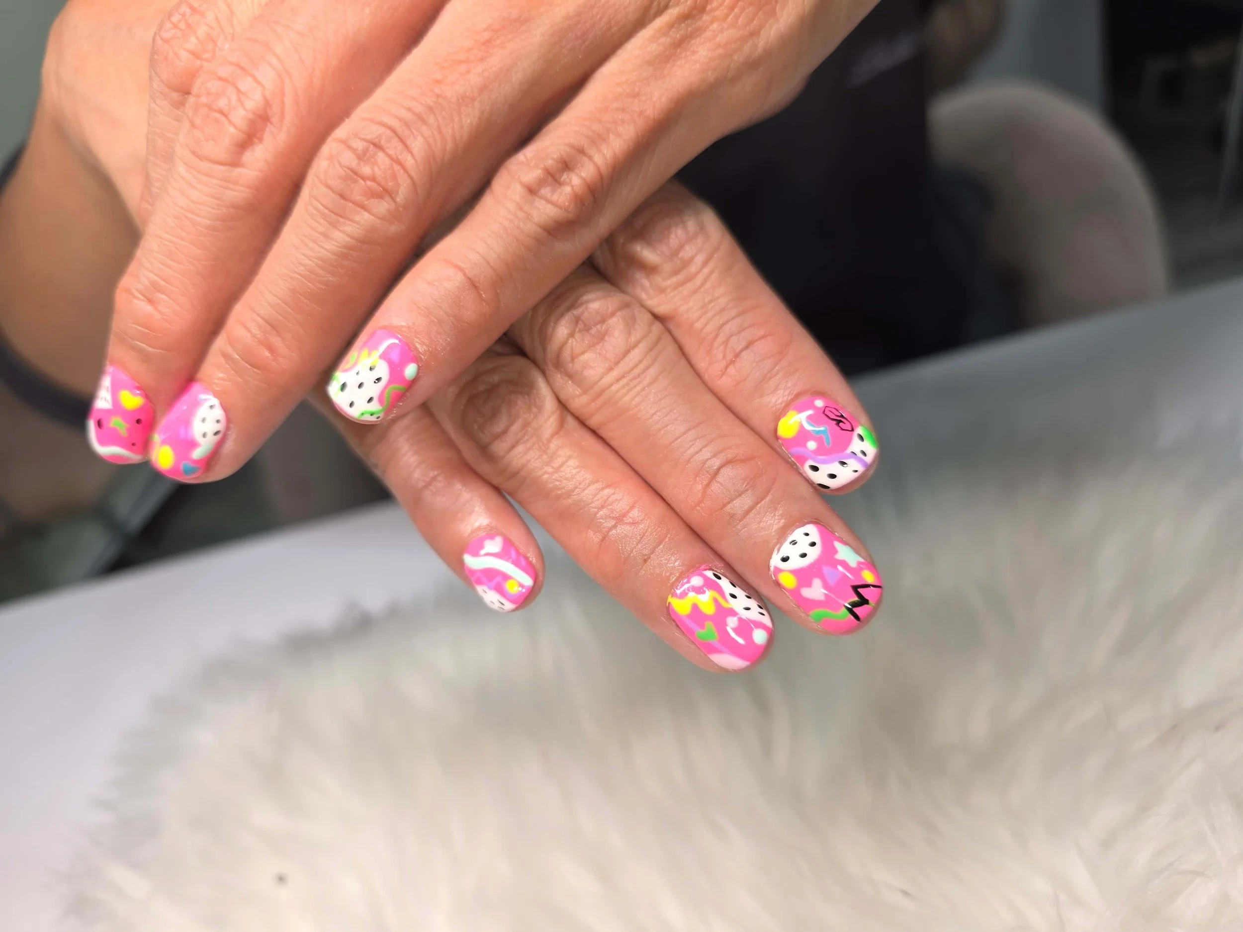 Close-up of hands with colorful, playful nail art featuring pink backgrounds with white, black, yellow, green, and blue accents in various patterns.