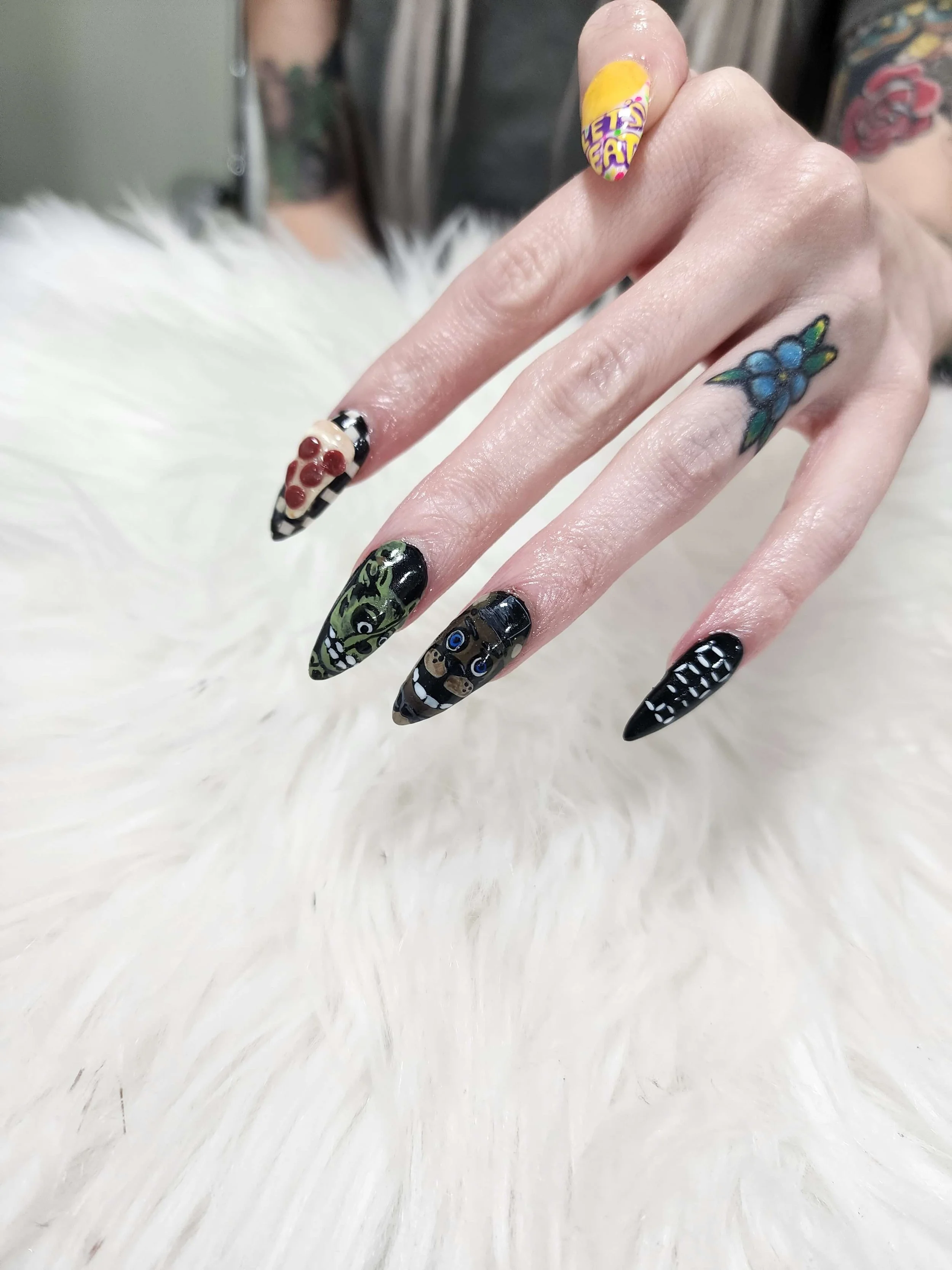 Close-up of a hand with decorated nails, each with unique black and other colored designs, including a green monster and a skull. The fingers are displayed against a white fluffy surface.