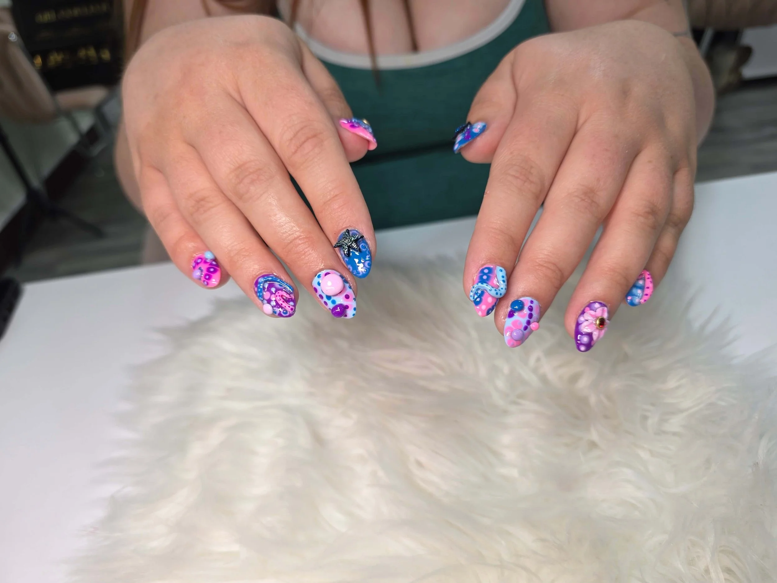 Close-up of a person's hands with colorful, decorated nails featuring pink, blue, purple, and black designs, with shimmering accents and 3D elements like dots and a starfish charm.
