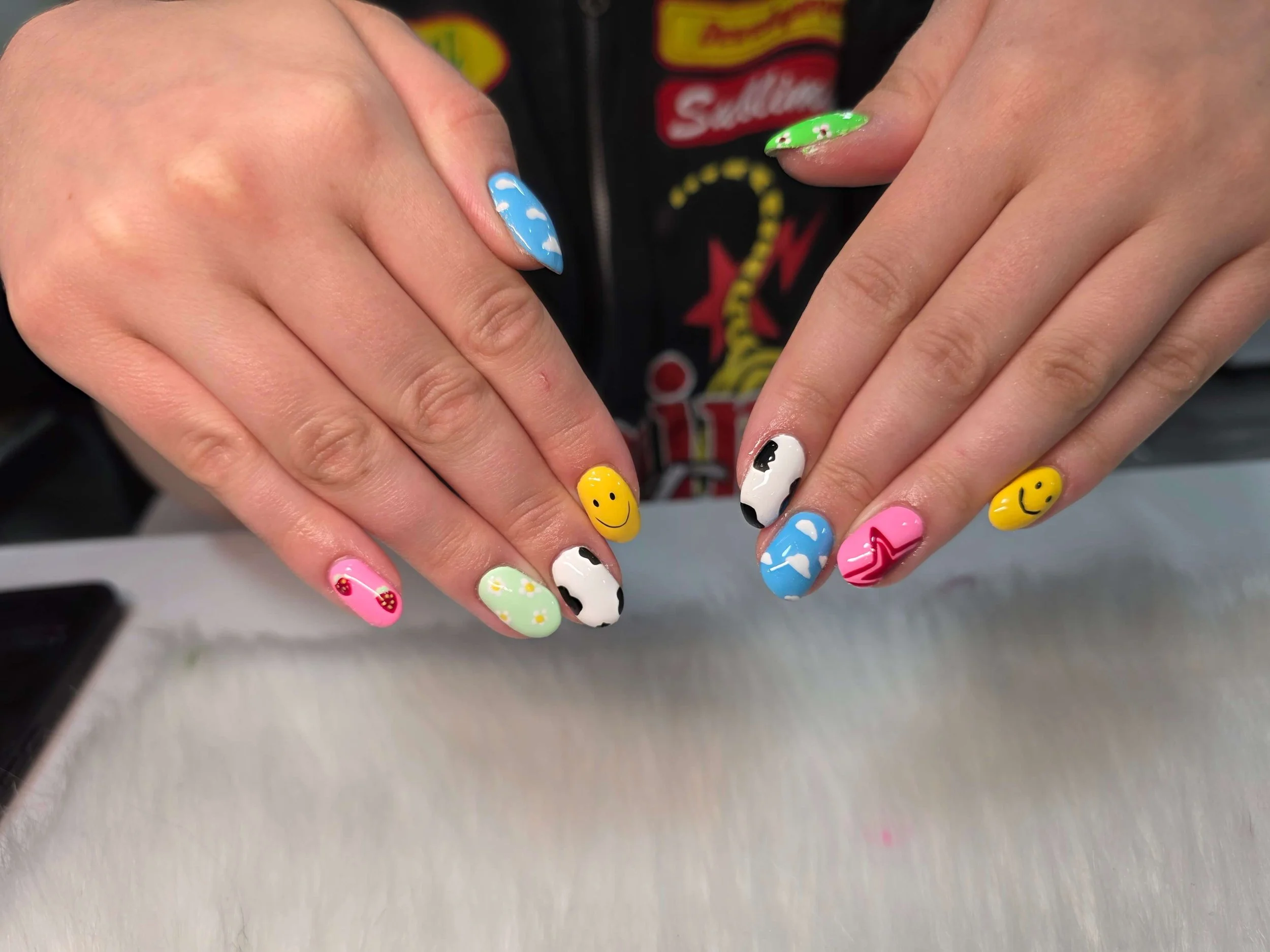 Close-up of a person's hands showing colorful nail art, including smiley faces, clouds, and abstract designs on each fingernail.