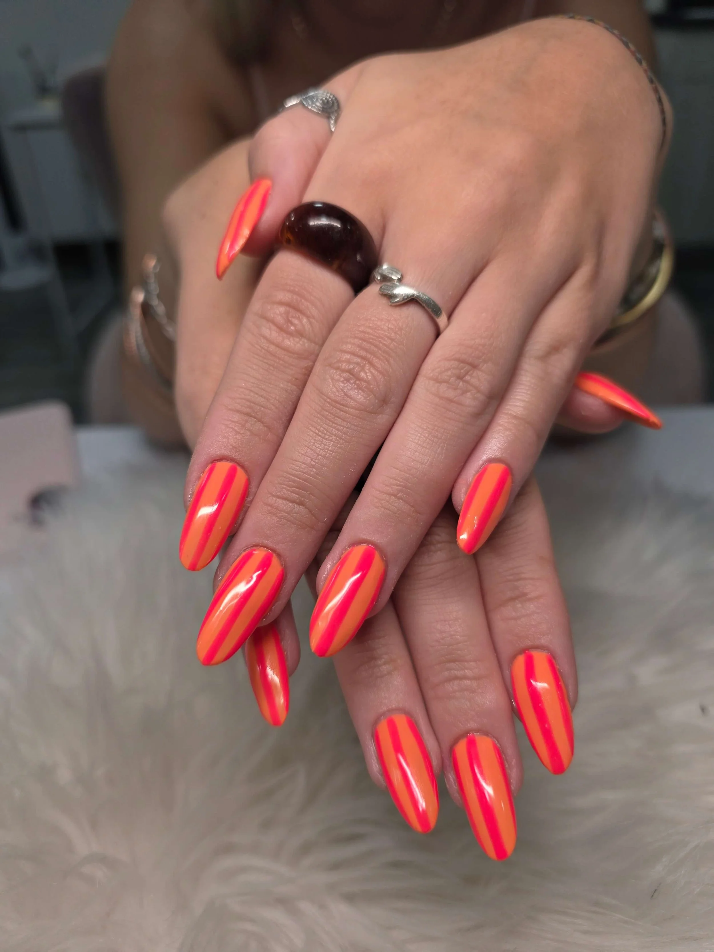 Close-up of a person's hands with long, neon orange and pink striped nails, wearing various rings, including one with a large dark gemstone, and another with a silver fish-shaped design. The background is blurred.