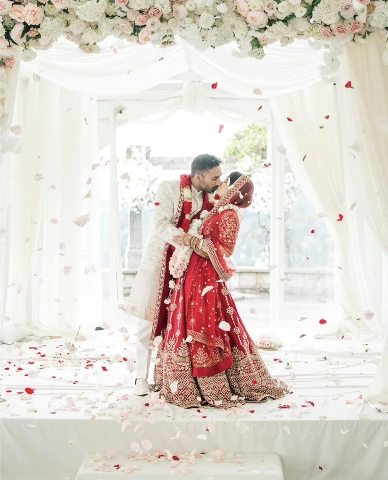 A couple dressed in traditional Indian wedding attire sharing a kiss under a decorated floral wedding canopy, with rose petals falling around them.