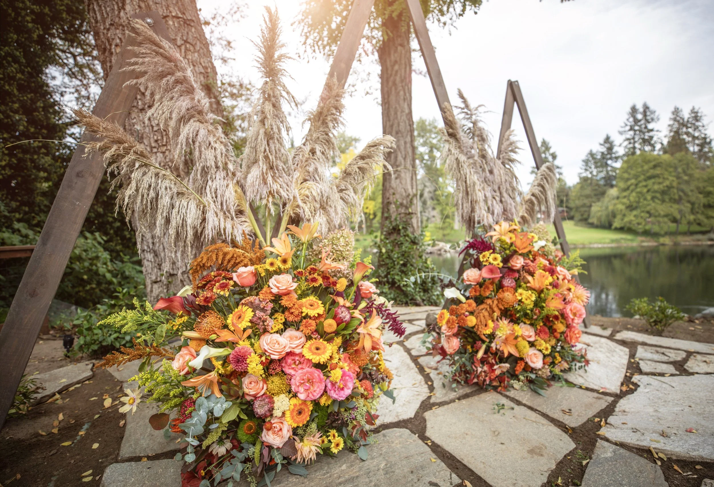 Colorful flower arrangements with pampas grass placed on stone pavement outdoors with trees and a river in the background.