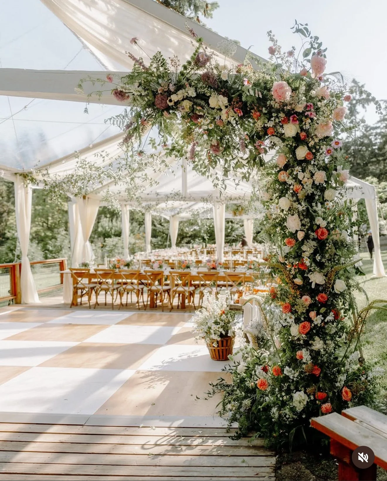 A decorated outdoor wedding reception area with a floral arch, long tables with chairs, and a white tent canopy. Flowers and greenery adorn the arch and surrounding area.