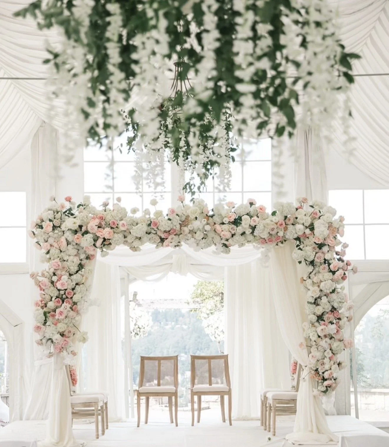 A decorated wedding arch with white and pink flowers in front of large windows inside a bright room.