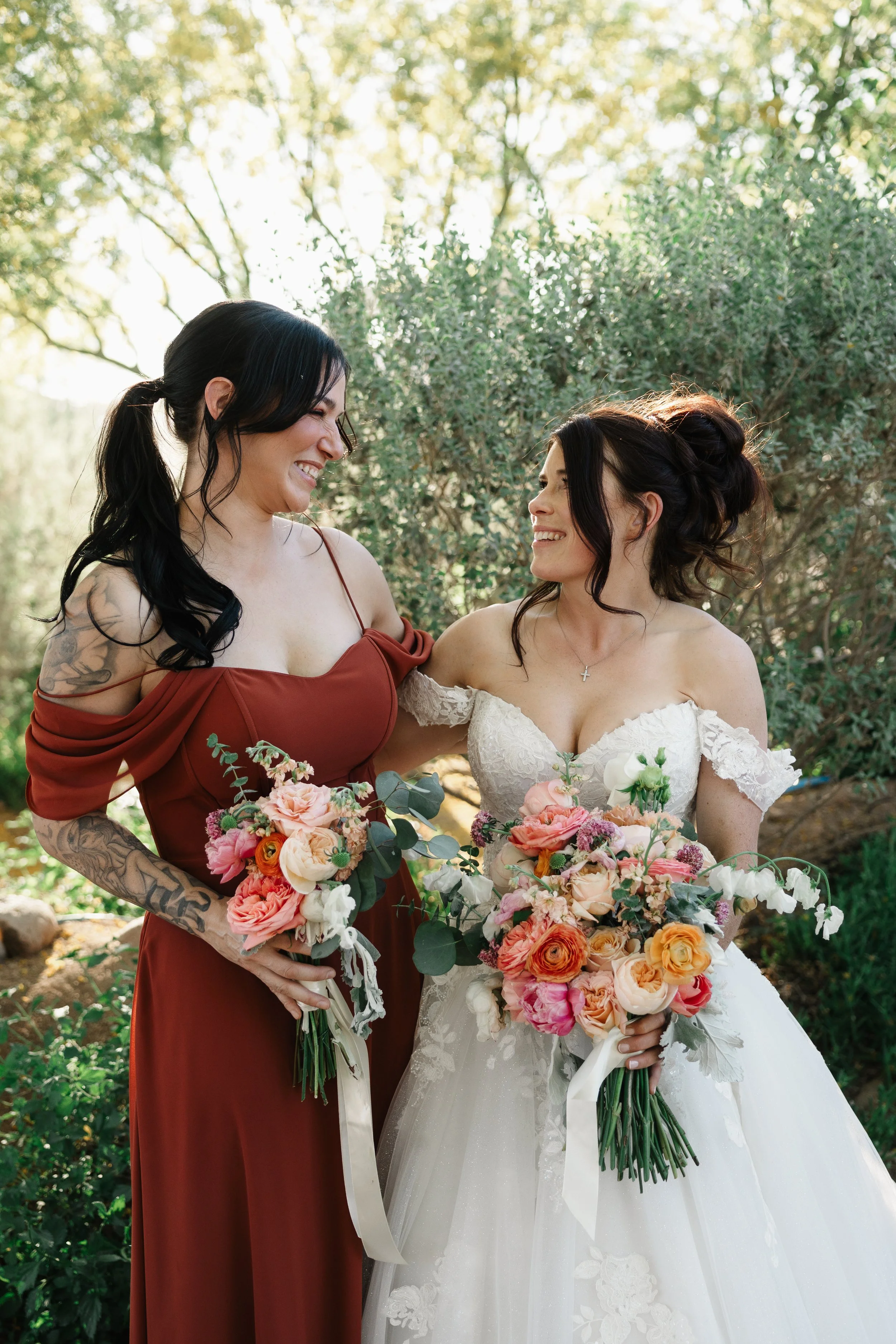 Two women, one in a bridal gown and the other in a red dress, smiling at each other while holding bouquets of flowers in an outdoor setting with green trees in the background.