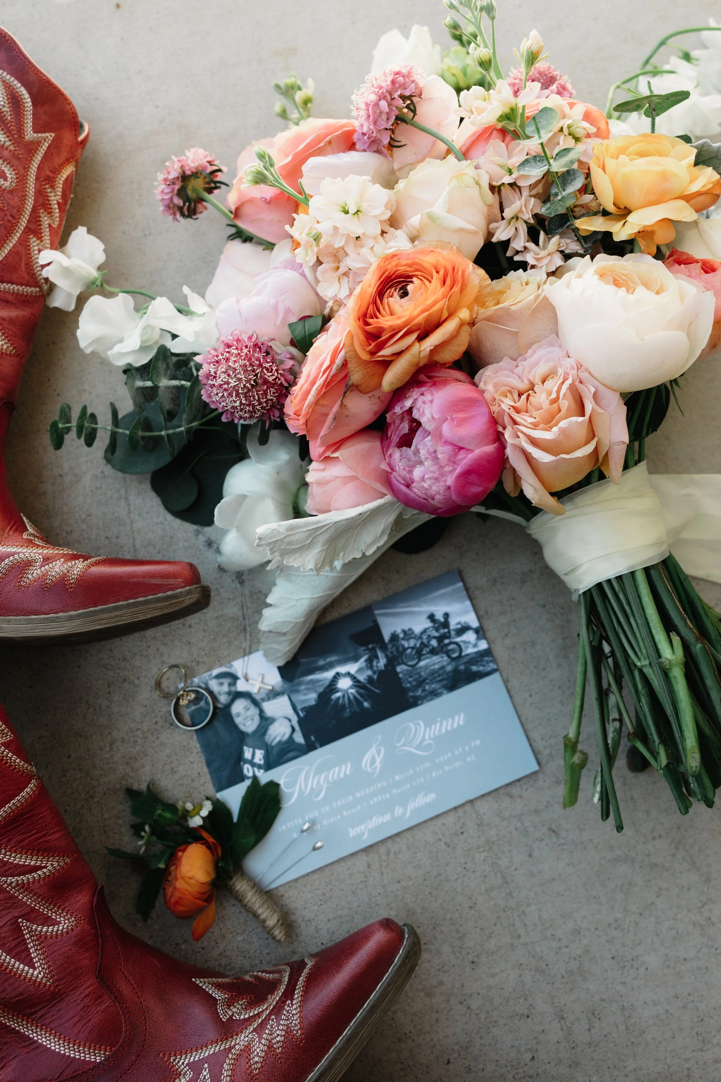 A bouquet of assorted flowers including roses, peonies, and other blooms in pastel shades of pink, peach, and white, resting on a light surface. Nearby are vintage red cowboy boots with decorative stitching, a black and white photo, wedding rings, and a wedding program for Megan and Quinn.