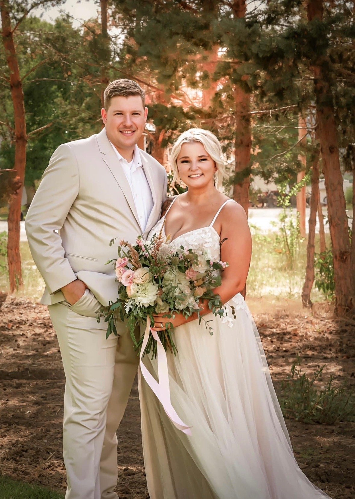 A newlywed couple standing outdoors among trees, the groom in a light beige suit and the bride in a white wedding gown holding a bouquet of flowers.
