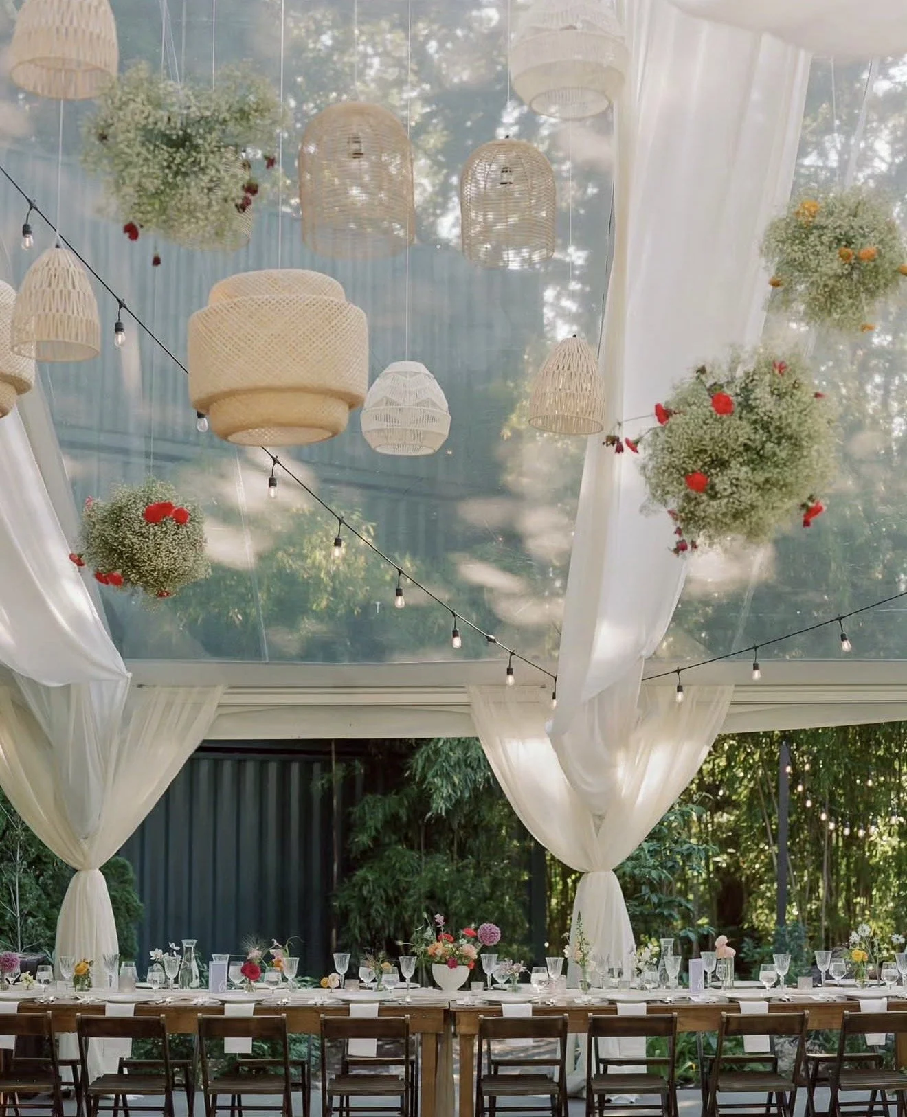 Decorated outdoor dining area under a glass roof with hanging wicker lanterns and floral arrangements, featuring tables with glassware and a white draped canopy.