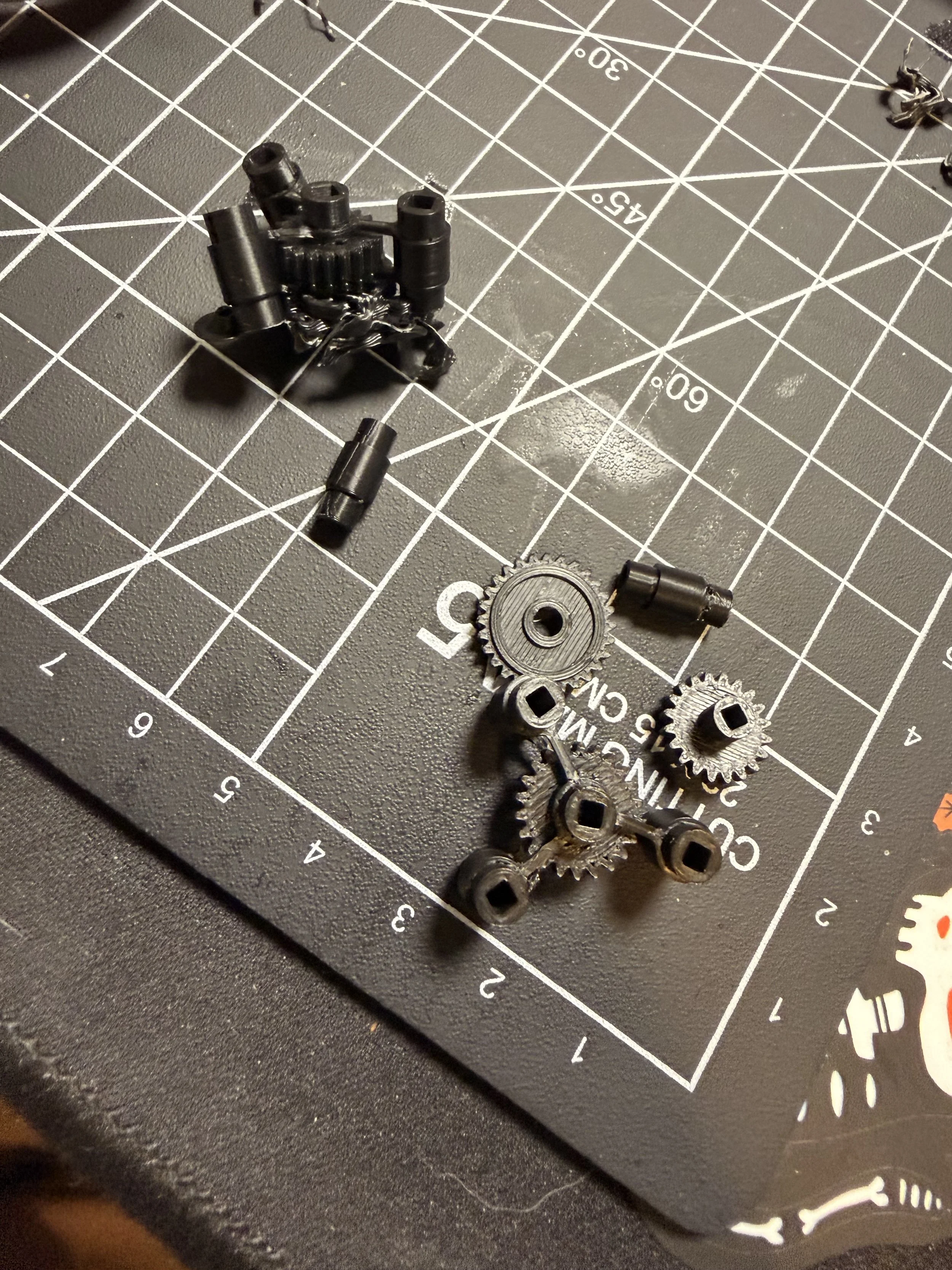 Small metal gears, screws, and pins placed on a black cutting mat with white measurement grid lines.