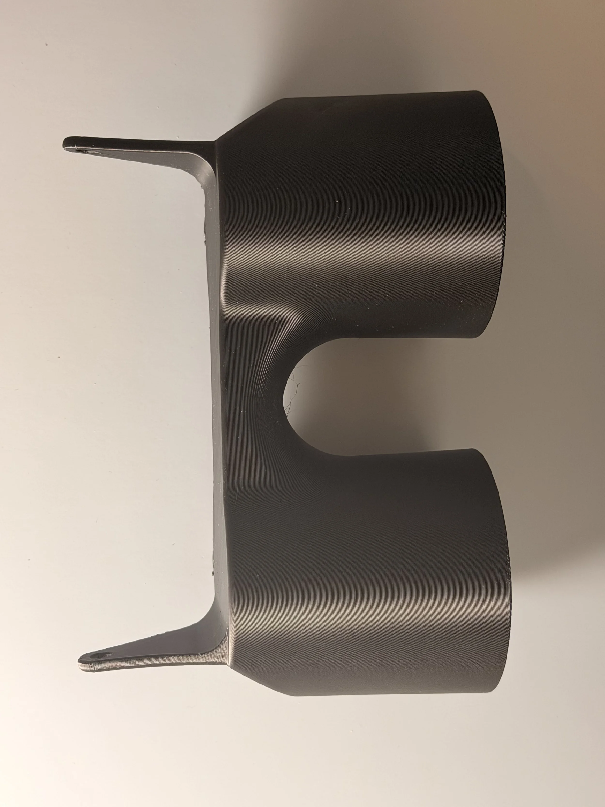 Close-up of a black metallic object with a flat, cylindrical shape and a tapered, elongated tip, possibly a headphone or a part of a device, placed on a white surface.