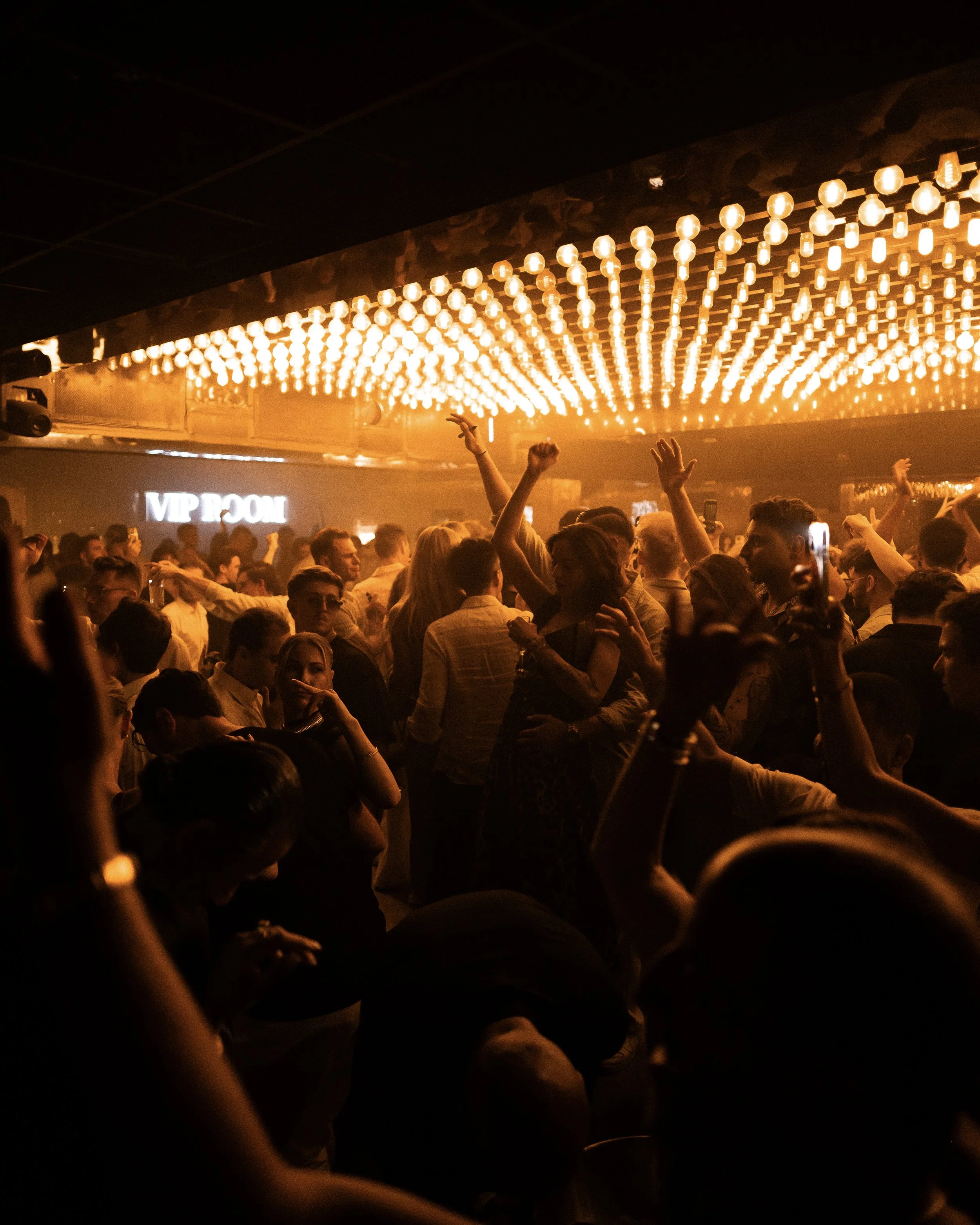 A crowded nightclub dance floor with people dancing and raising their hands, illuminated by a large, glowing, pattern of warm yellow-orange lights on the ceiling.