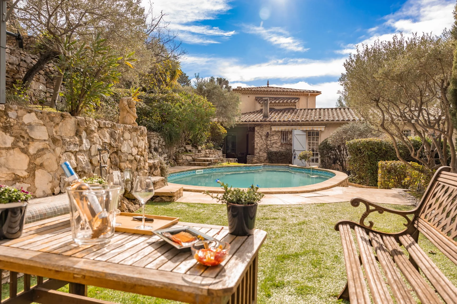 A backyard patio scene with a round swimming pool, surrounded by trees and a stone wall. There is outdoor furniture including a wooden table with drinks and snacks, and a bench. The sky is partly cloudy with sunlight.