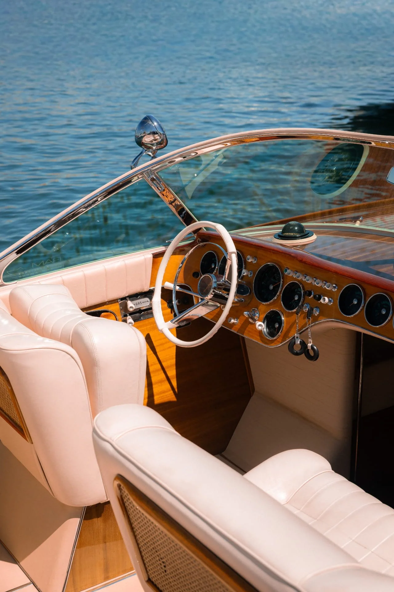 The image shows the interior of a vintage boat with a wooden dashboard, white leather seats, and a view of water outside through a windshield.