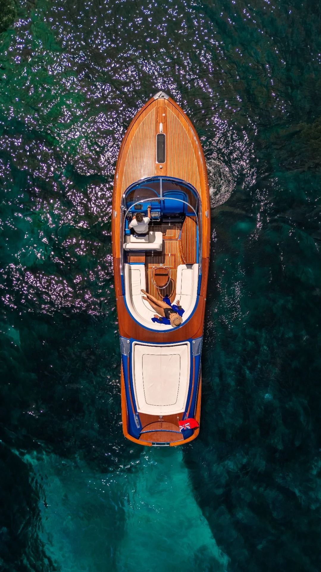 Overhead view of a wooden boat with two people on board, surrounded by clear blue water with sunlight reflections.