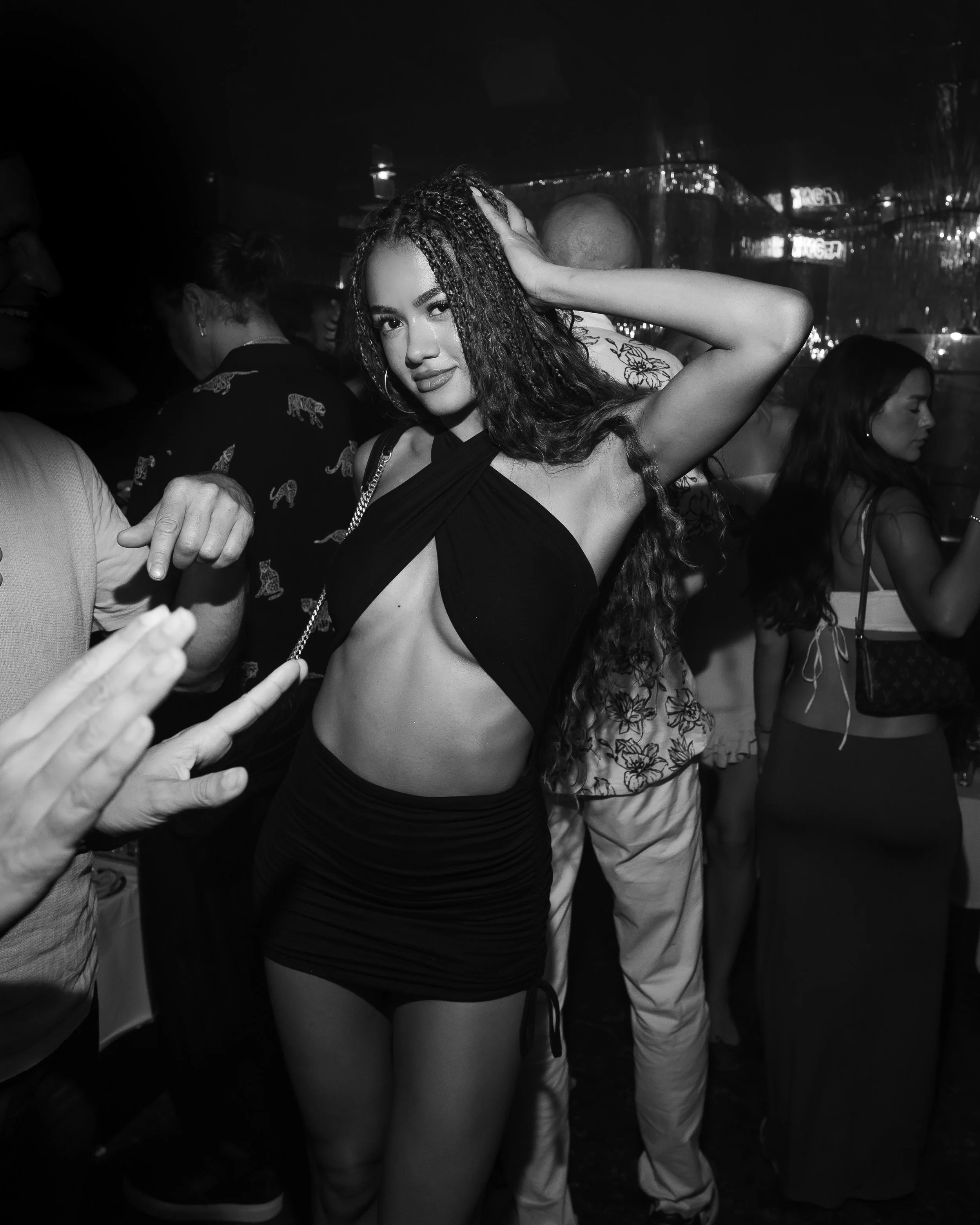 A woman with long, curly hair wearing a black, sleeveless top and black pants poses in a crowded nightclub with her hand behind her head, surrounded by other people, in a black-and-white photo.