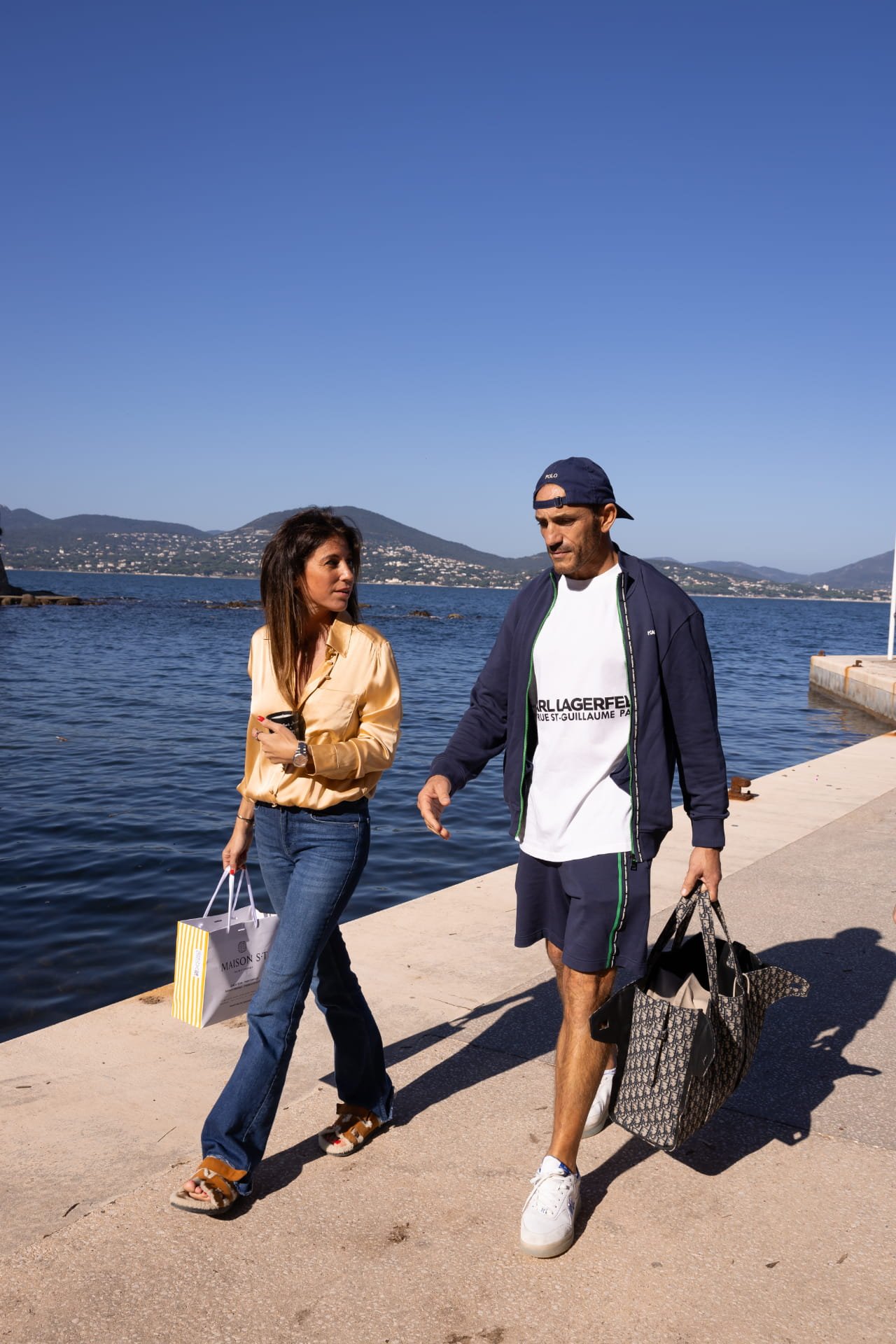 A man and woman walking along a waterfront promenade, carrying shopping bags. The woman has long dark hair, wearing a gold blouse, blue jeans, and sandals. The man has a beard, wearing a blue cap, navy jacket, white T-shirt, shorts, and white sneaker