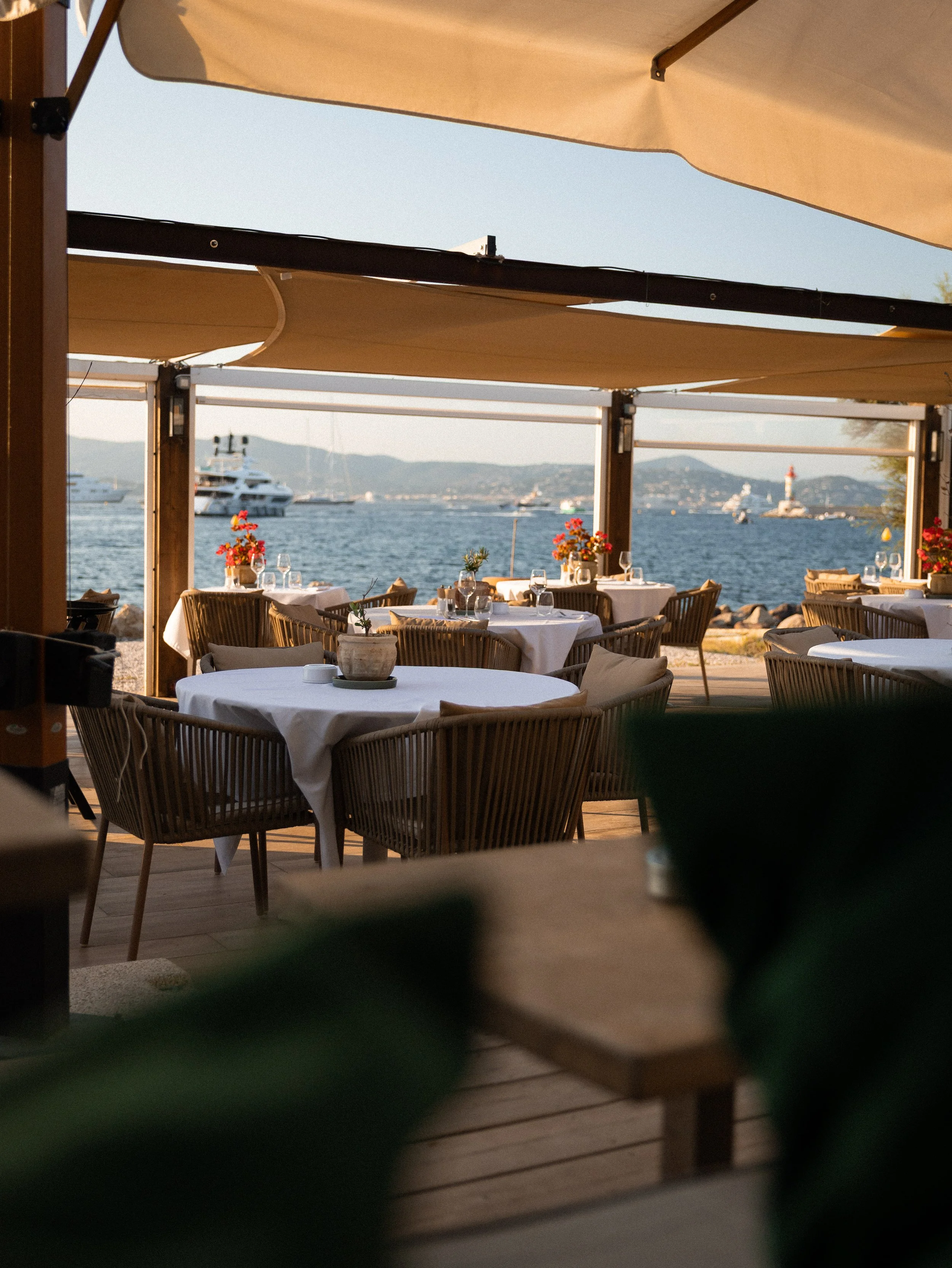 Outdoor restaurant with tables set with white tablecloths, glasses, and flowers, overlooking a body of water with boats and a lighthouse in the distance, shaded by beige sun umbrellas.