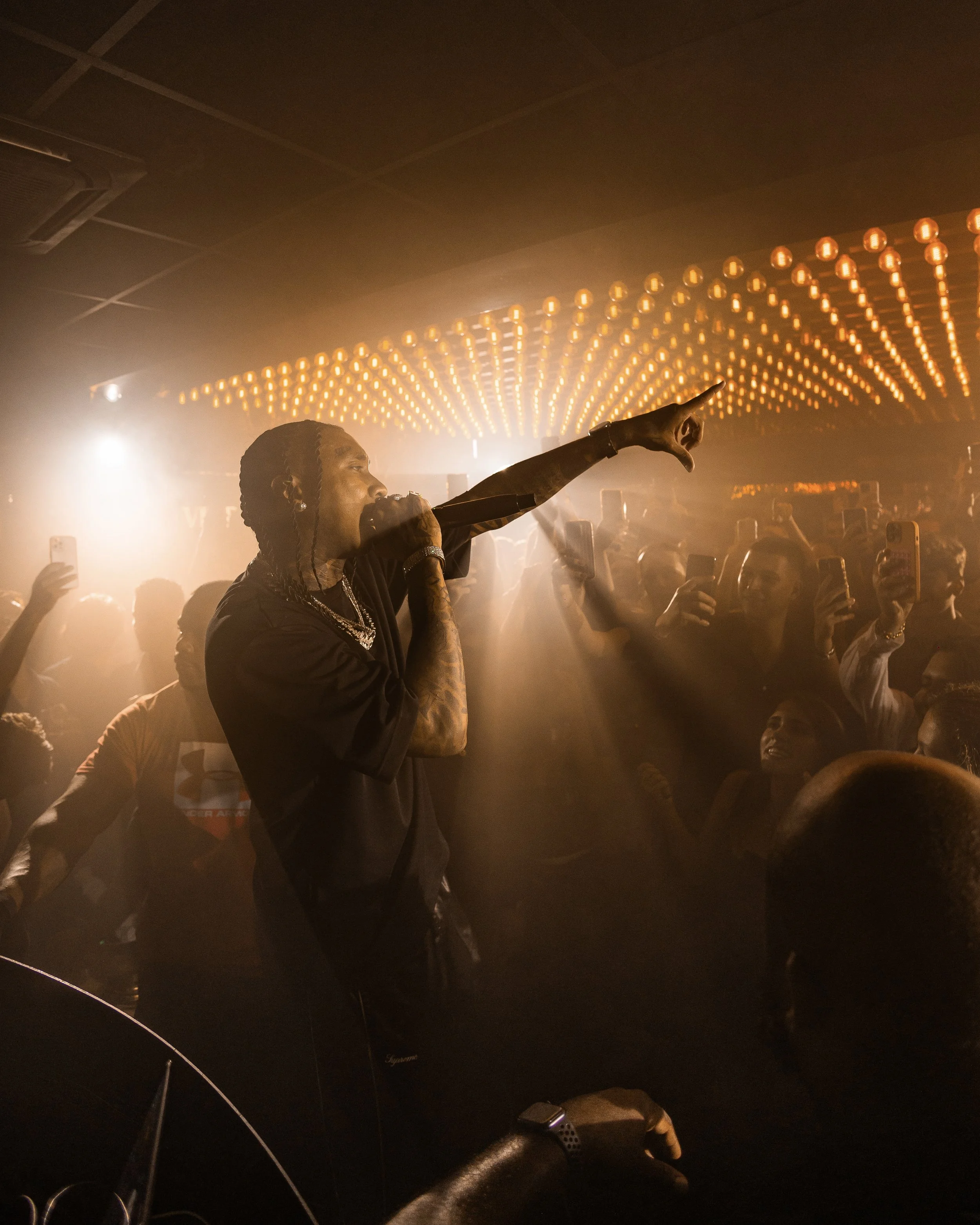 A person holding a microphone on stage with a crowd of people in front, illuminated by orange lights on the ceiling, creating a warm atmosphere.