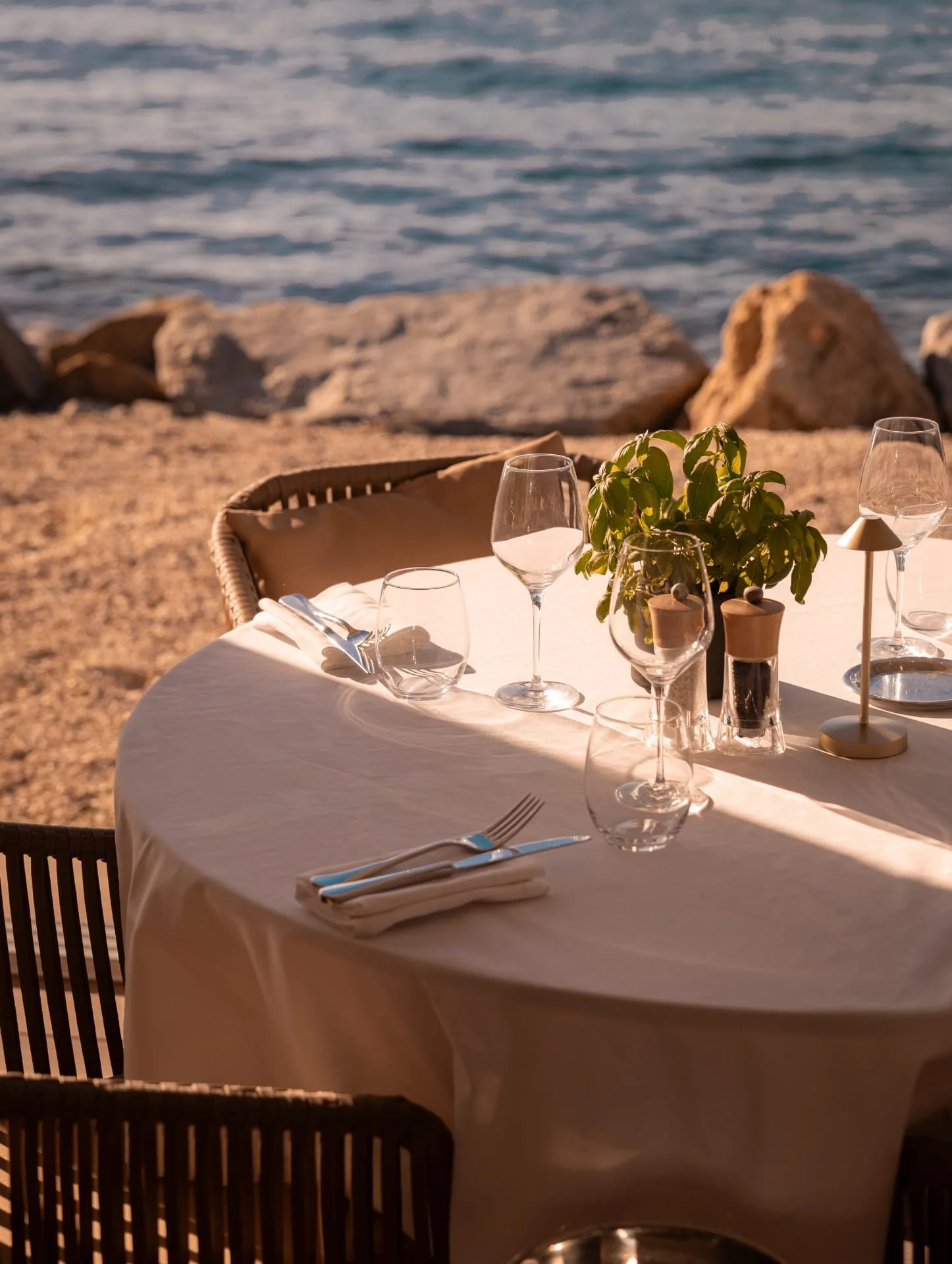 An outdoor dining table set with glassware, cutlery, and a potted plant, overlooking a rocky shoreline and the water.
