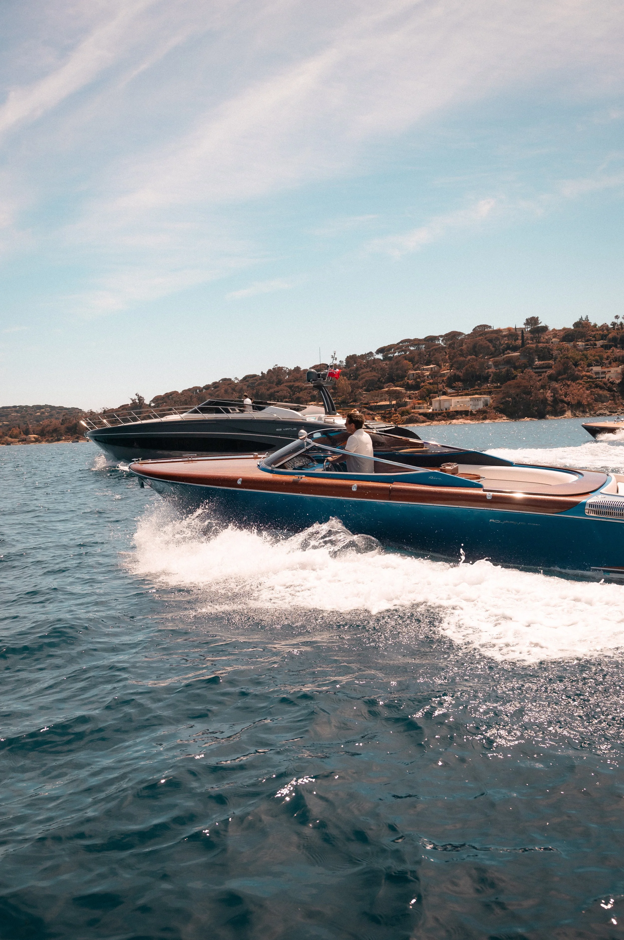 Two speedboats speeding on the water with a hilly shoreline and houses in the background under a partly cloudy sky.