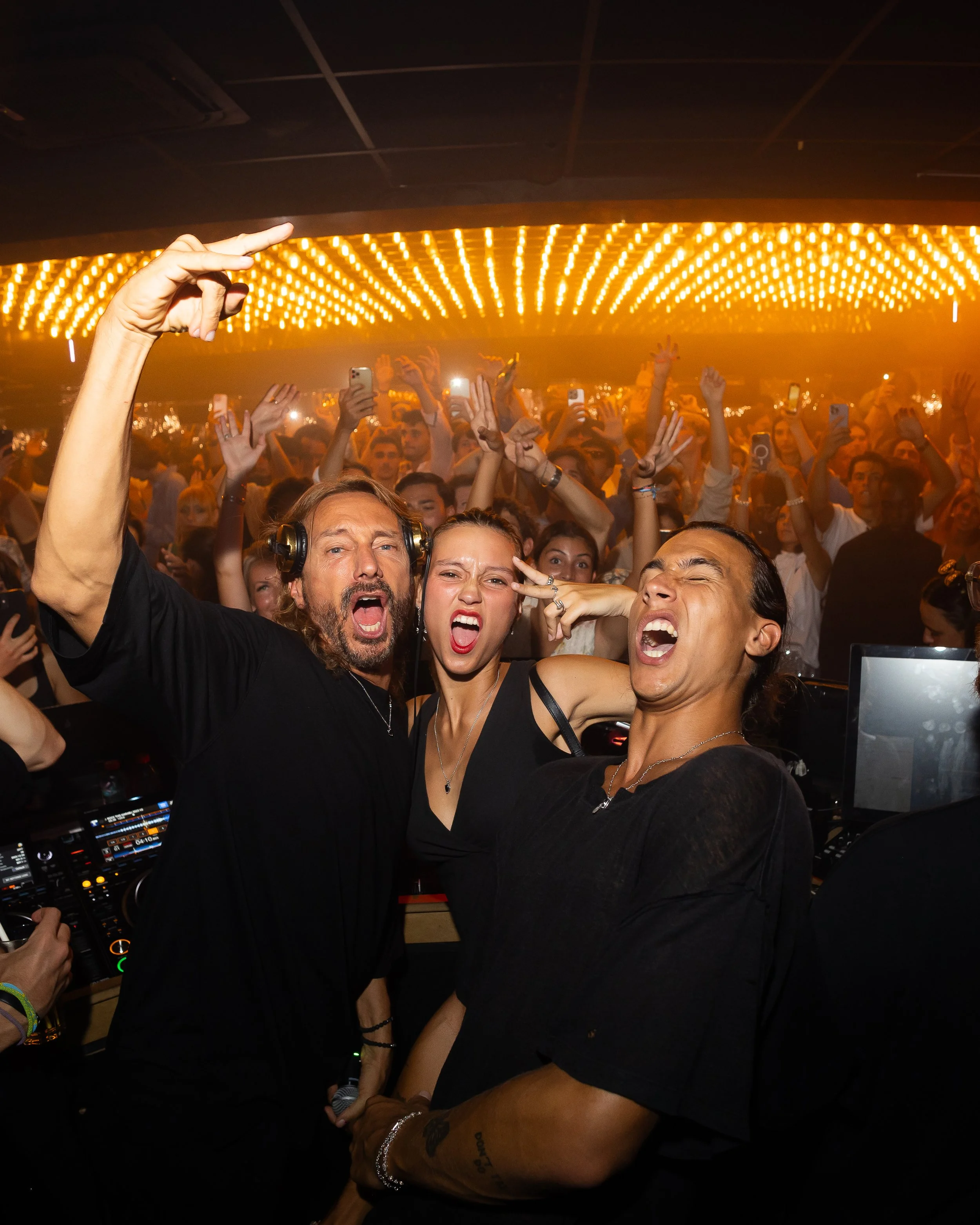 Crowd of people dancing and taking photos at a nightclub with a DJ booth and a yellow LED ceiling display.