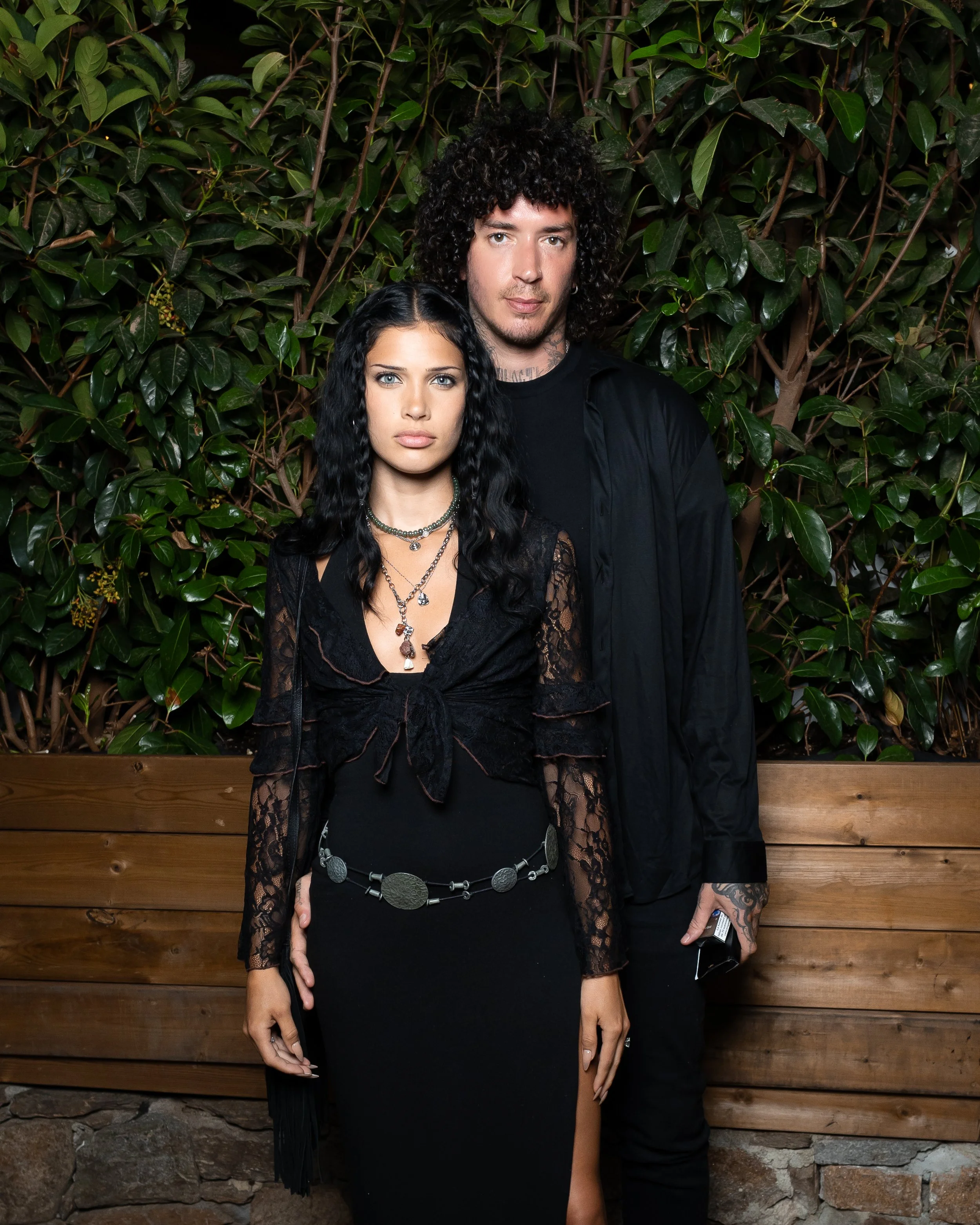 A man and woman standing in front of a leafy green wall and a wooden fence. The woman has long black wavy hair, light skin, and is wearing a black lace dress with layered necklaces. The man has curly dark hair, light skin, and is wearing a black shir