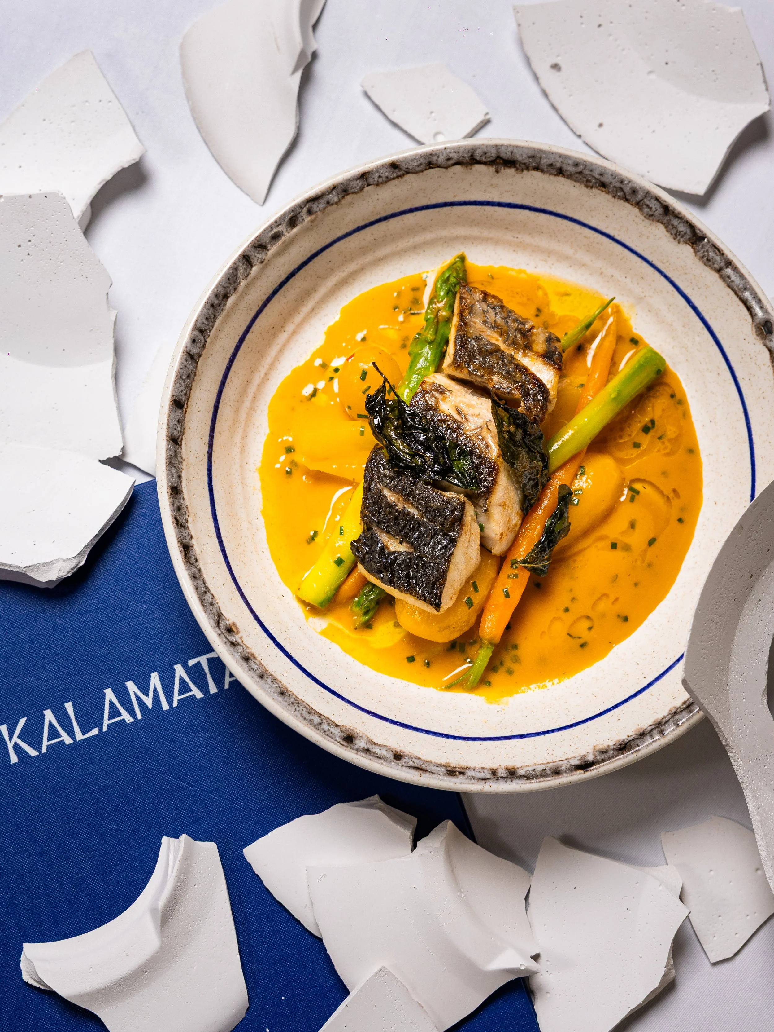 A plate of cooked fish with crispy skin, served with green beans, carrots, and yellow sauce, surrounded by broken white plates and a blue mat.
