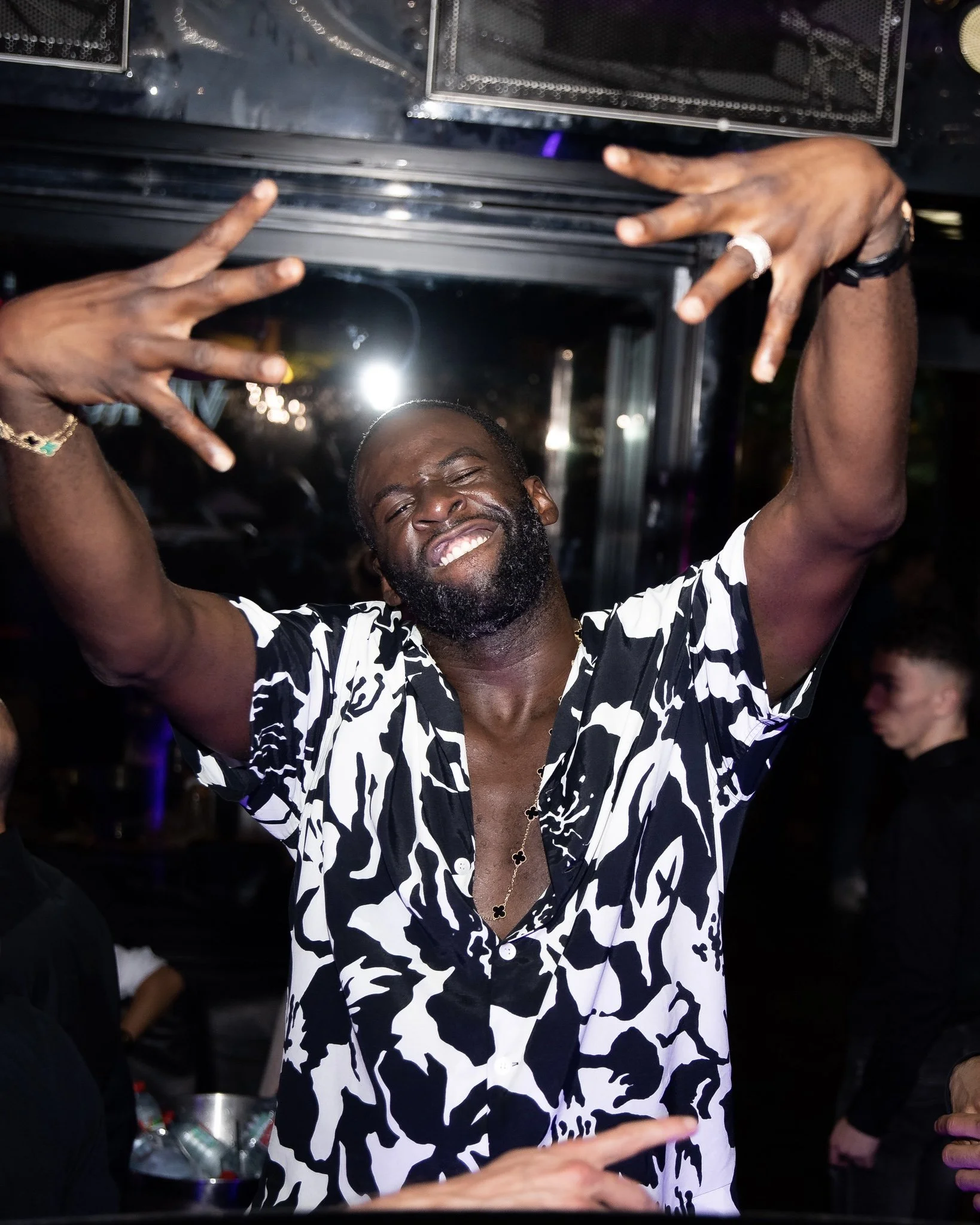 A man smiling and making hand gestures with both hands in a lively, dark indoor setting, possibly a nightclub or party, with other people in the background and bottles on a table.