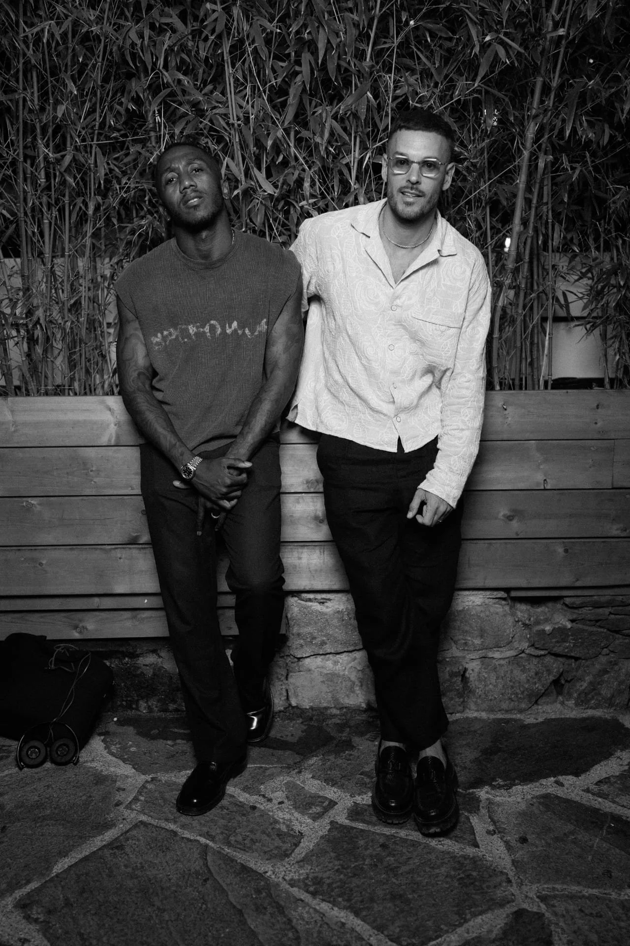 Two men standing side by side against a wooden fence and bamboo plants. The man on the left has dark skin, a sleeveless shirt, and tattoos on his arms. The man on the right has light skin, glasses, and is wearing a patterned button-up shirt. Both are