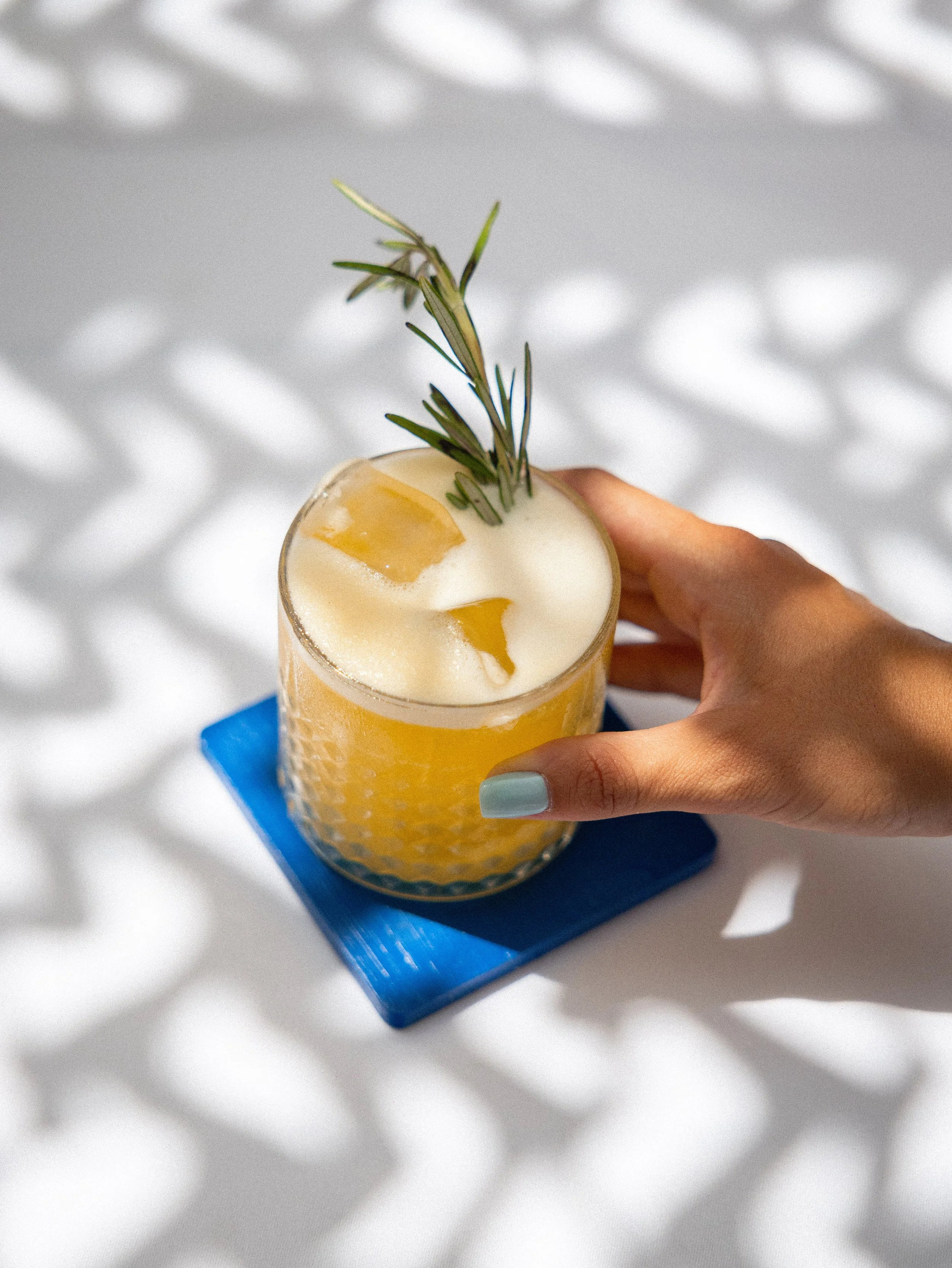 Hand holding a glass of yellow beverage, garnished with a sprig of rosemary and lemon slices, placed on a blue coaster on a white surface.