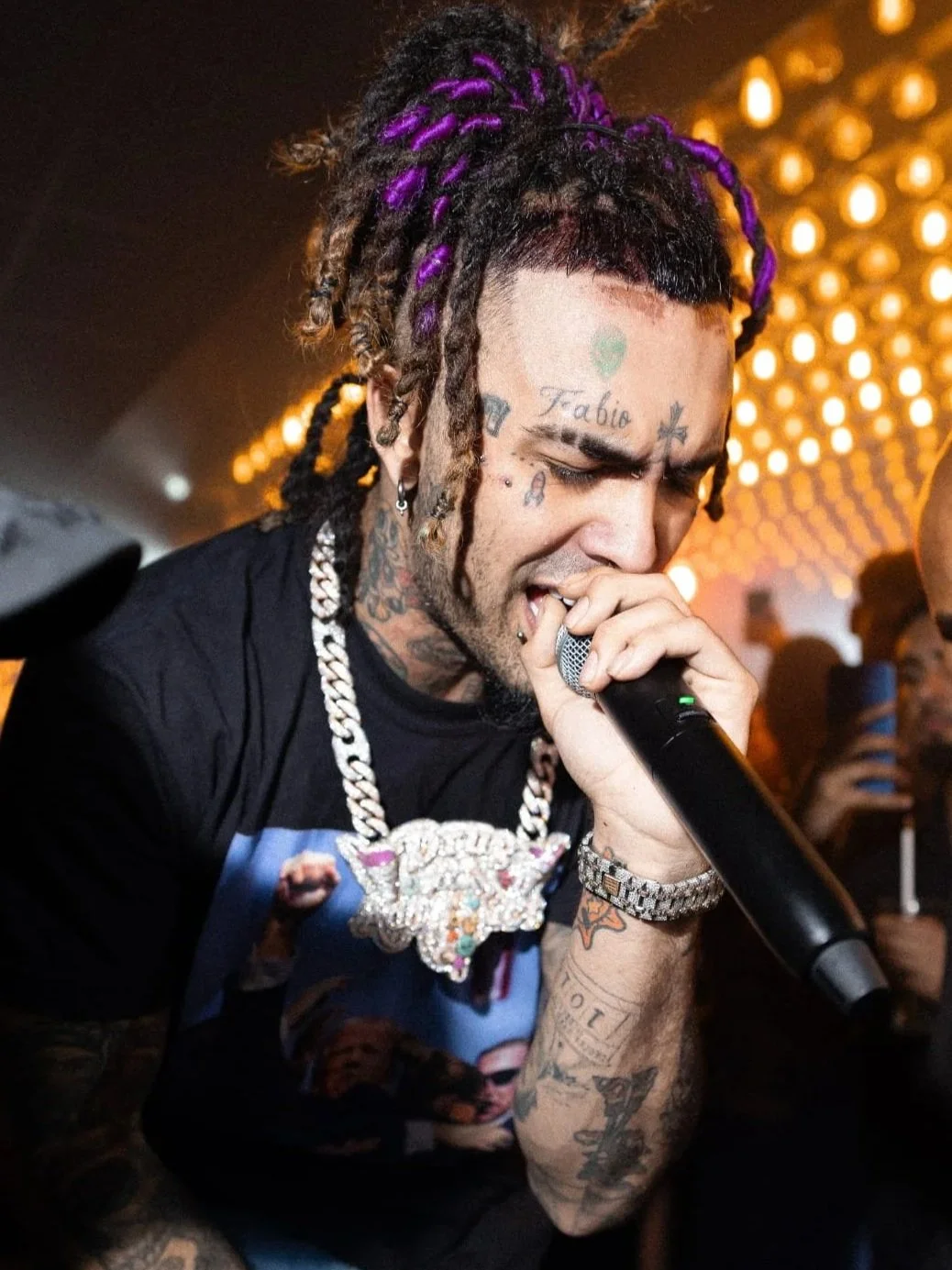 A man with tattoos and purple dreadlocks holding a microphone, singing passionately in a nightclub with a glowing, dotted ceiling in the background.