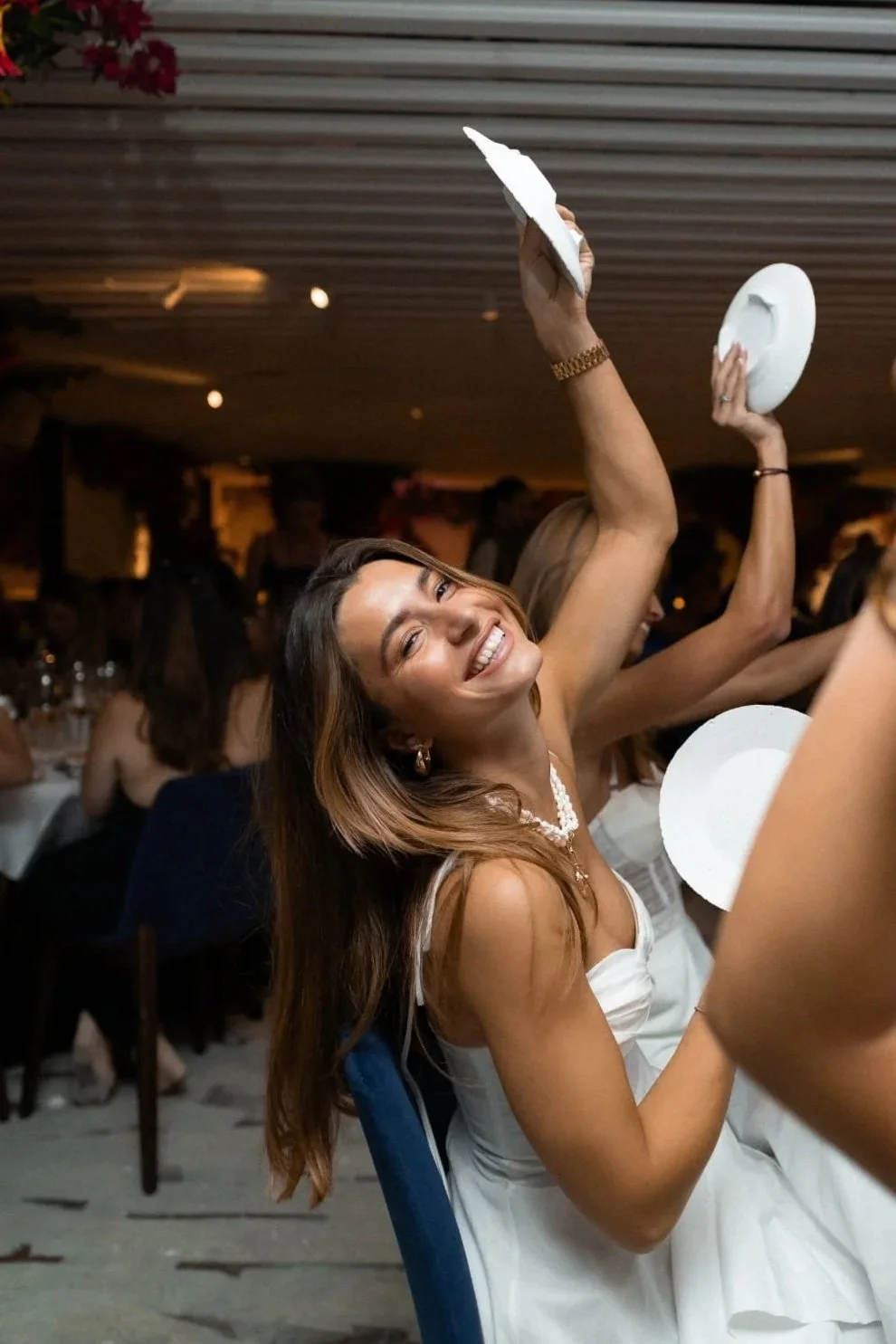 A woman smiling and enjoying herself at a social gathering, sitting at a table with people in the background, holding a plate with her hand raised.
