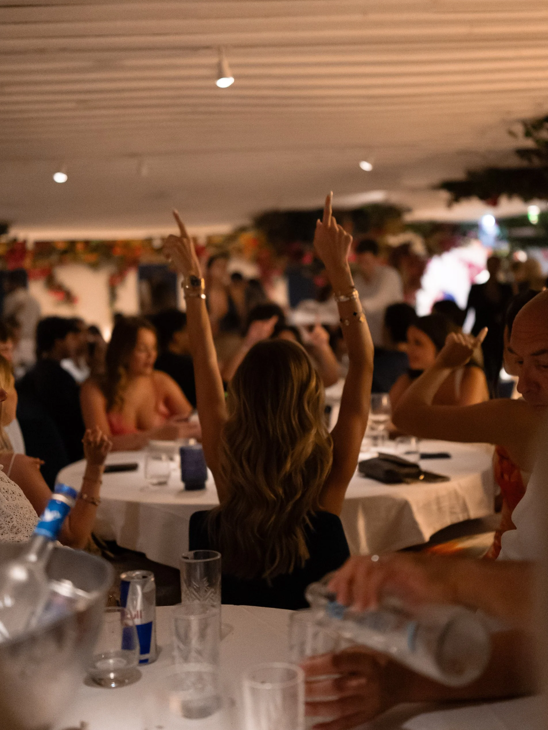 A crowded indoor event with people seated at tables, some raising their hands, decorated with garlands and flowers, with a focus on a woman with long hair raising her hand.