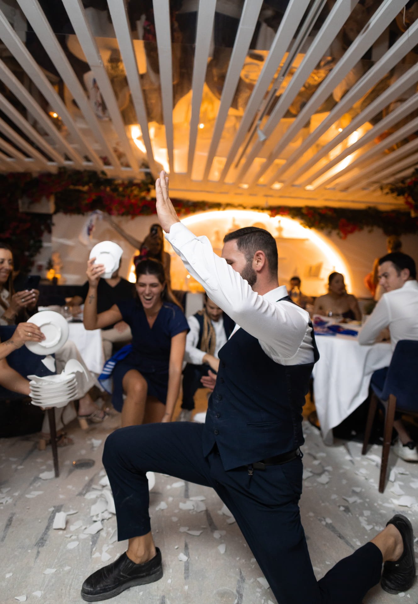 A man dressed in a formal vest and pants kneeling on one knee while holding his hand up, surrounded by a group of people in a decorated indoor setting. Some are taking photos and others are smiling, with celebratory paper scattered on the floor and t