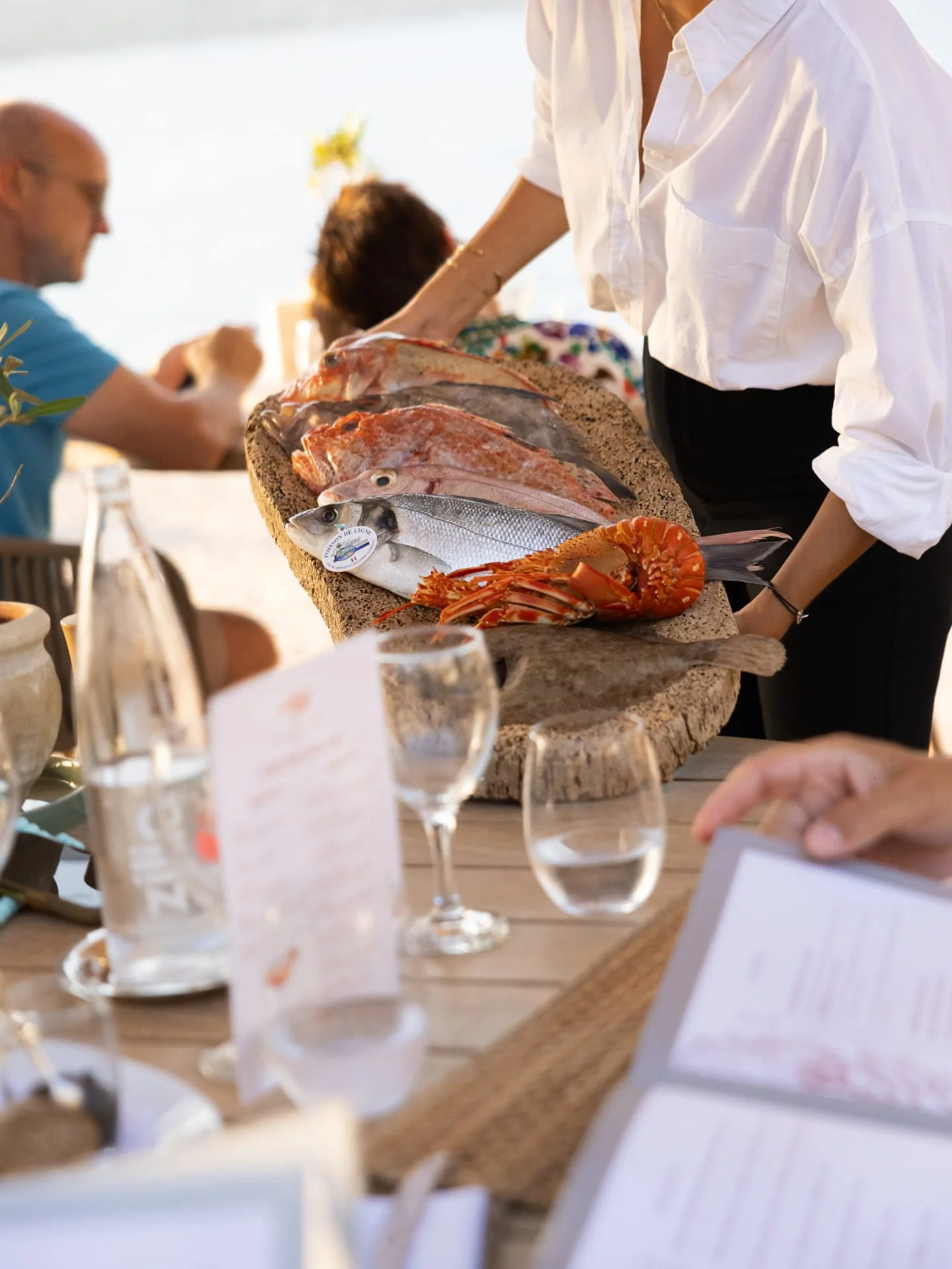 A person in a white shirt arranging fresh seafood, including fish, lobster, and other shellfish, on a large wooden serving board at a gathering or meal.