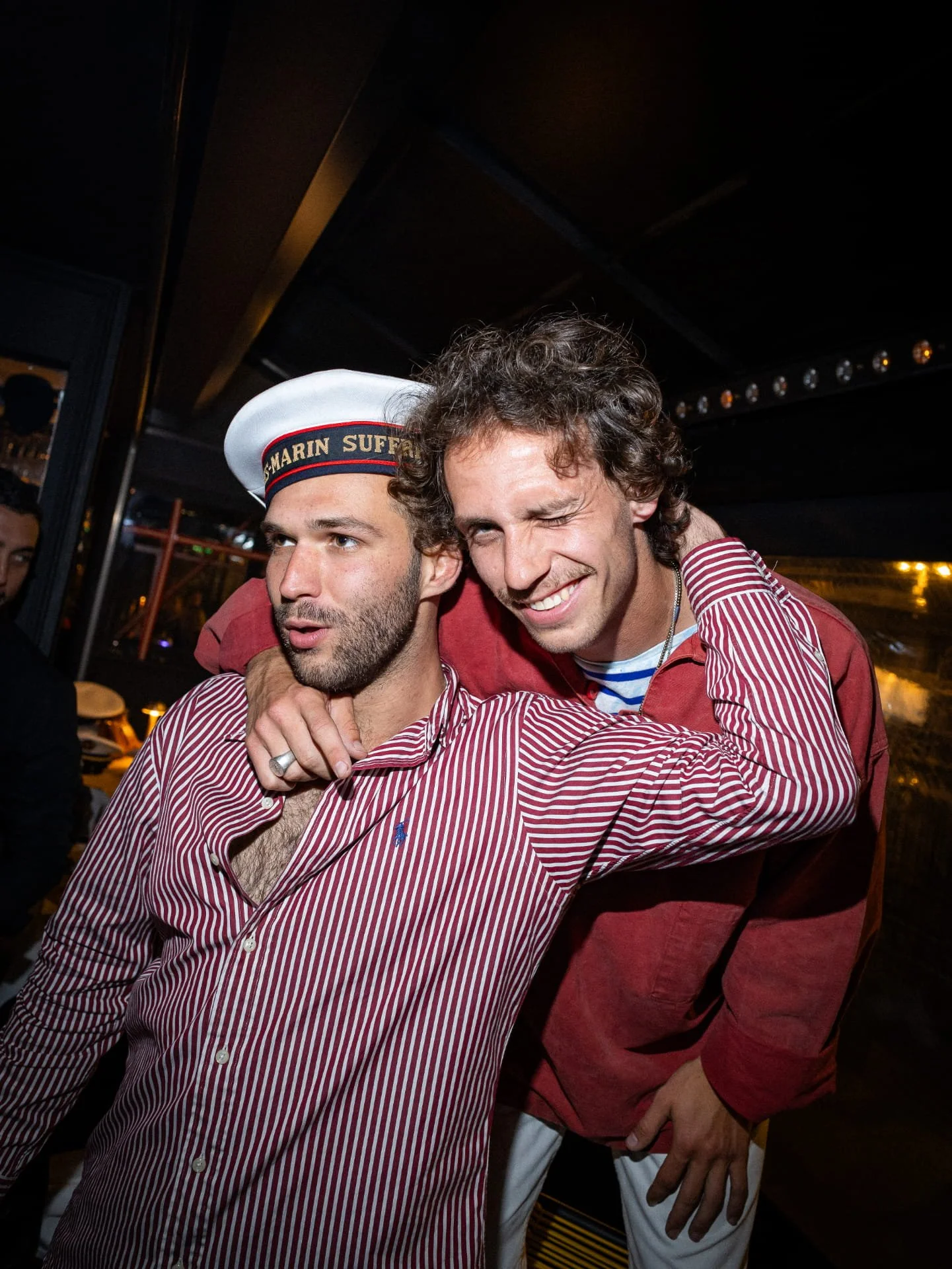 Two men celebrate at a party, one wearing a white sailor hat with black and red band that reads 'Sail Marin Surf', and the other with curly hair and a big smile, wearing a red jacket over a striped shirt. They are hugging and one is winking while the