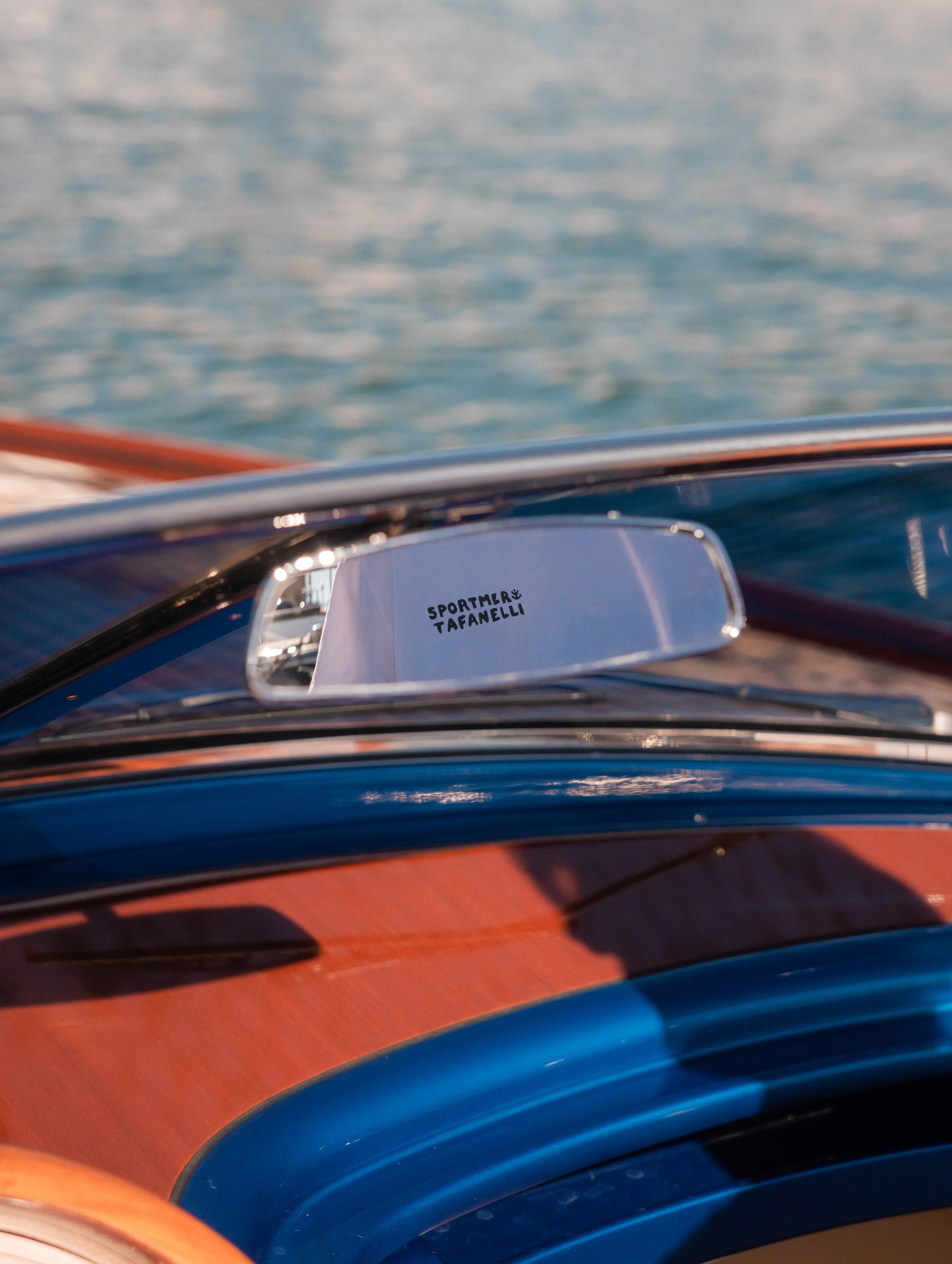A mirror on a vehicle reflecting a person wearing a white shirt with black text 'SPORTMERY TAFANELLI' and colorful stripes on the vehicle's blue and orange body, with a background of water.