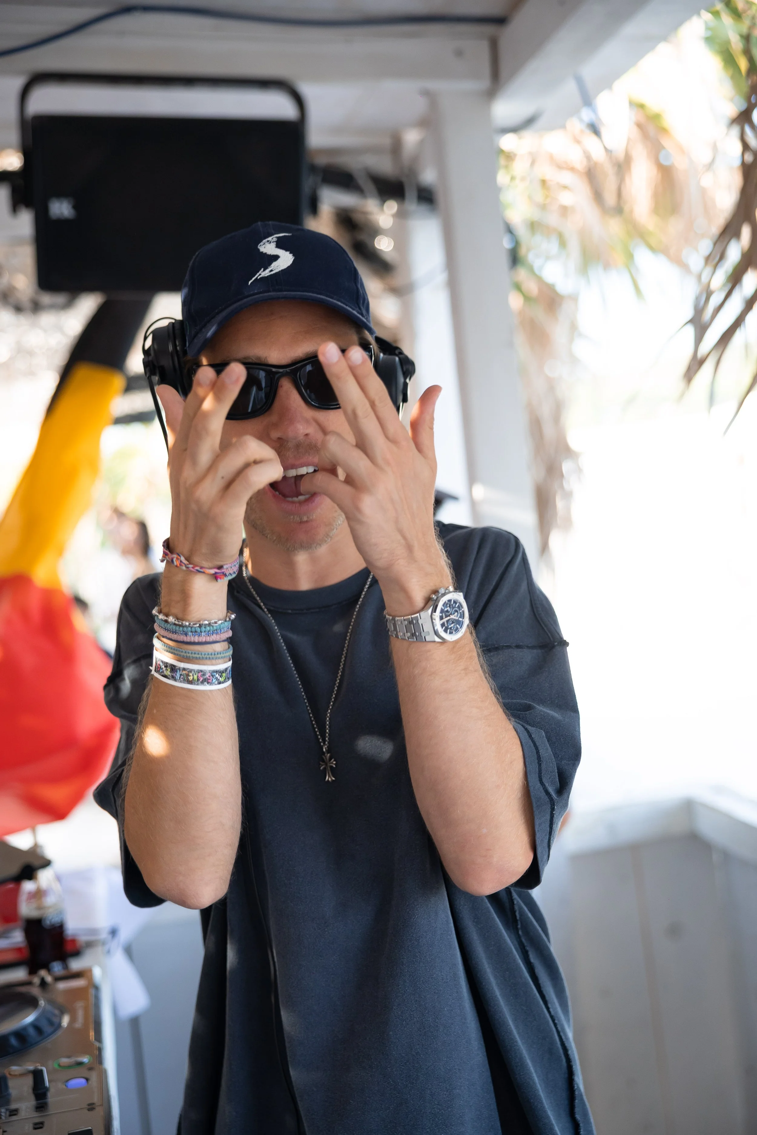 Young man wearing sunglasses, a navy cap, and multiple bracelets on his wrist, adjusting his sunglasses in an outdoor setting with sunlight and foliage in the background.