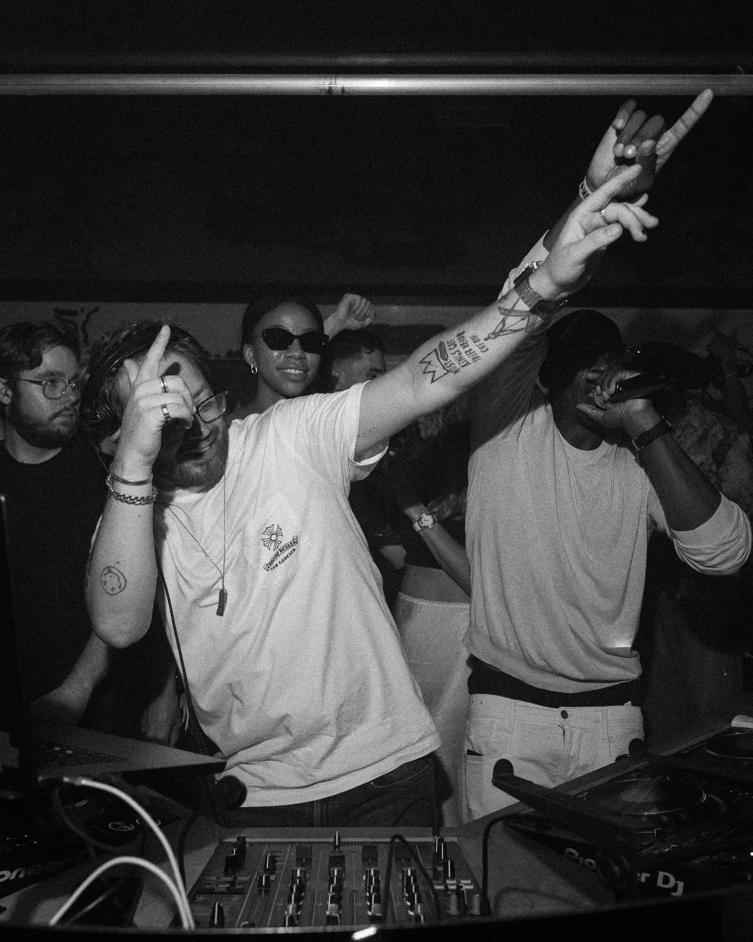 People dancing and DJ equipment in a dark club setting with a woman smiling and making a peace sign, and a man singing into a microphone.