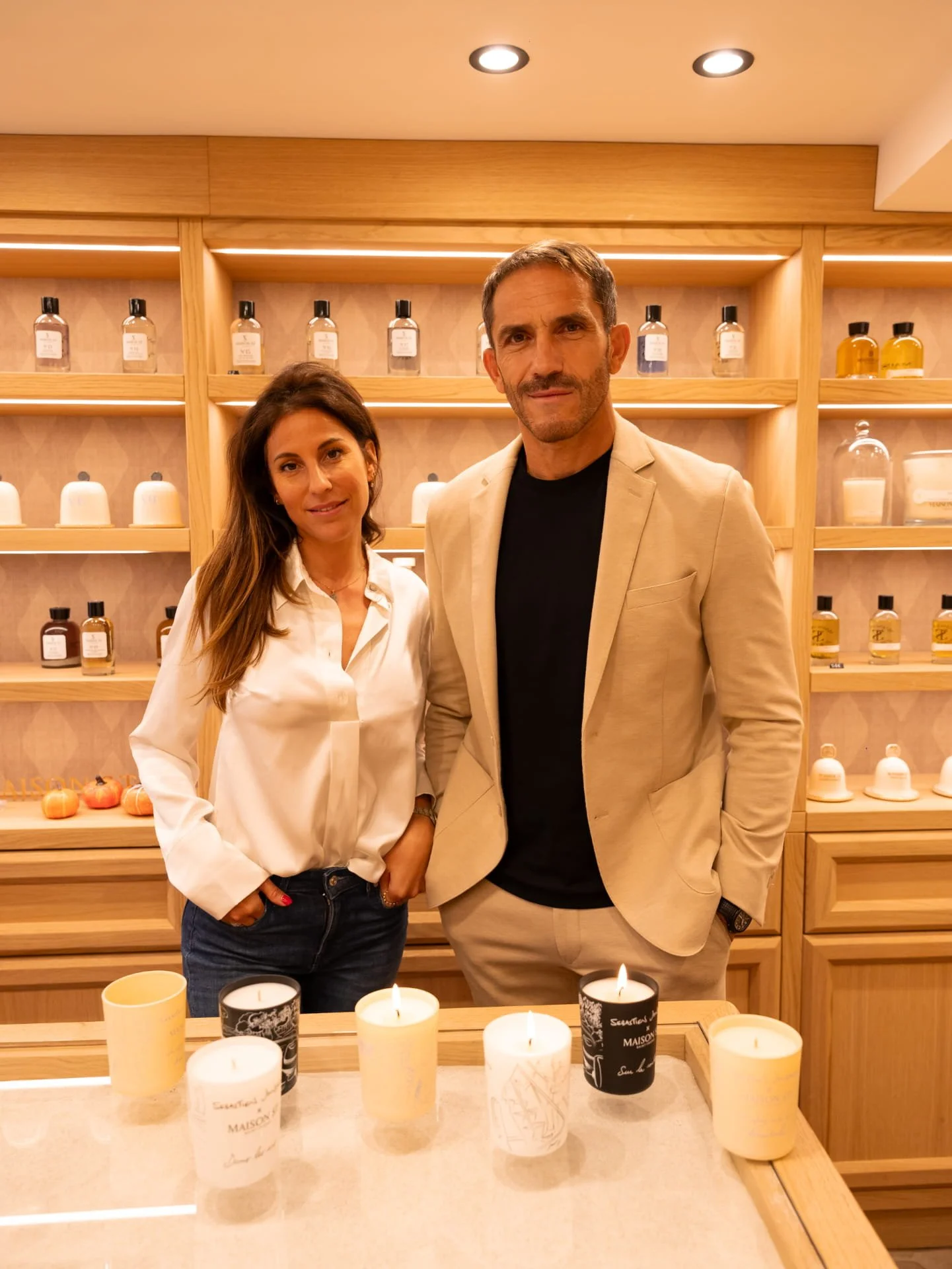 A man and a woman standing together in a room with wooden shelves behind them displaying bottles and ceramics. Candles are on the table in front of them.
