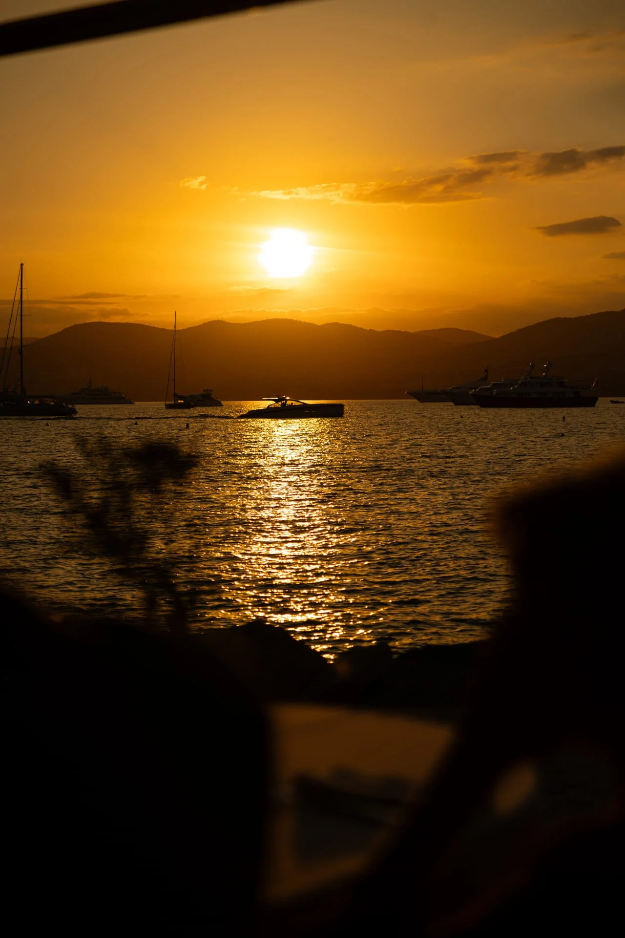 Sunset over a body of water with boats and yachts, mountains in the background, and trees in the foreground.